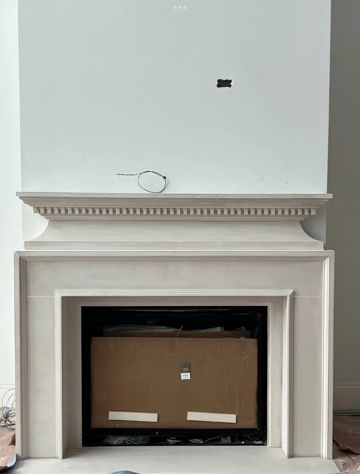 Unfinished fireplace with a decorative mantel and a cardboard box inside the fireplace opening, with some electrical wiring and a hole in the wall above.