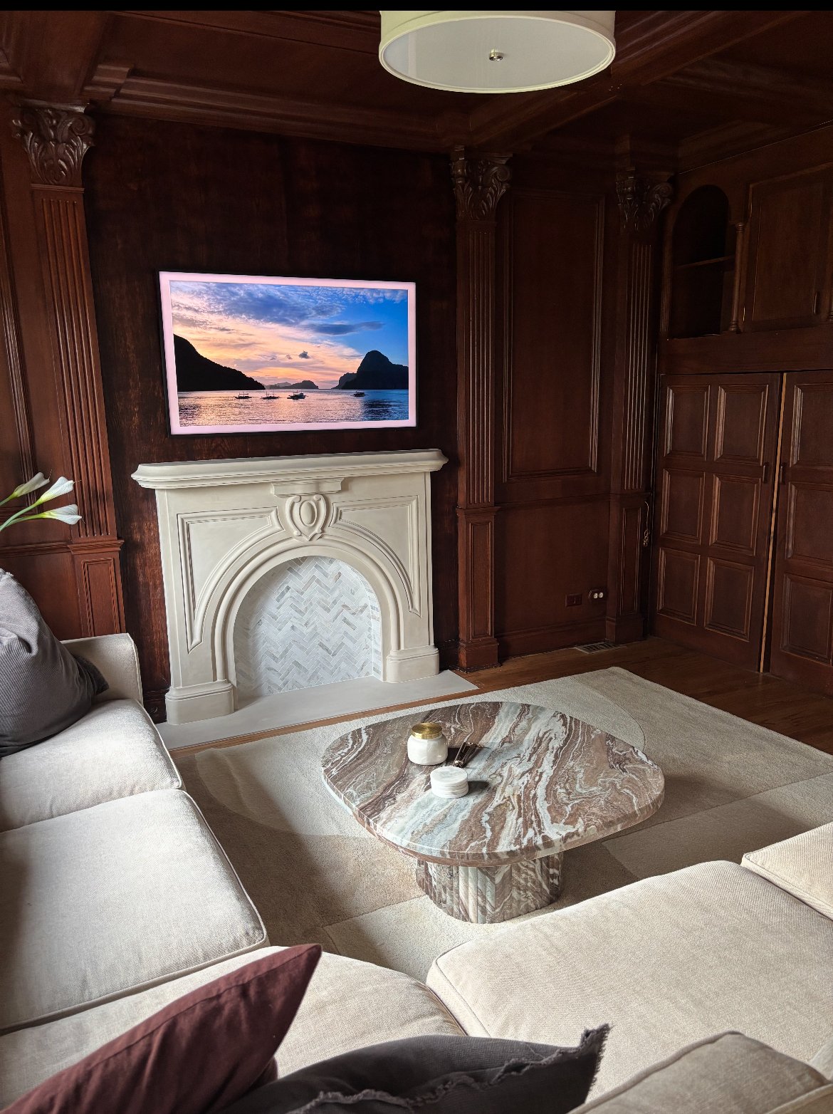 Living room with dark wooden paneling, cream-colored fireplace, large television displaying a seascape, marble coffee table, and beige sofas with pillows.