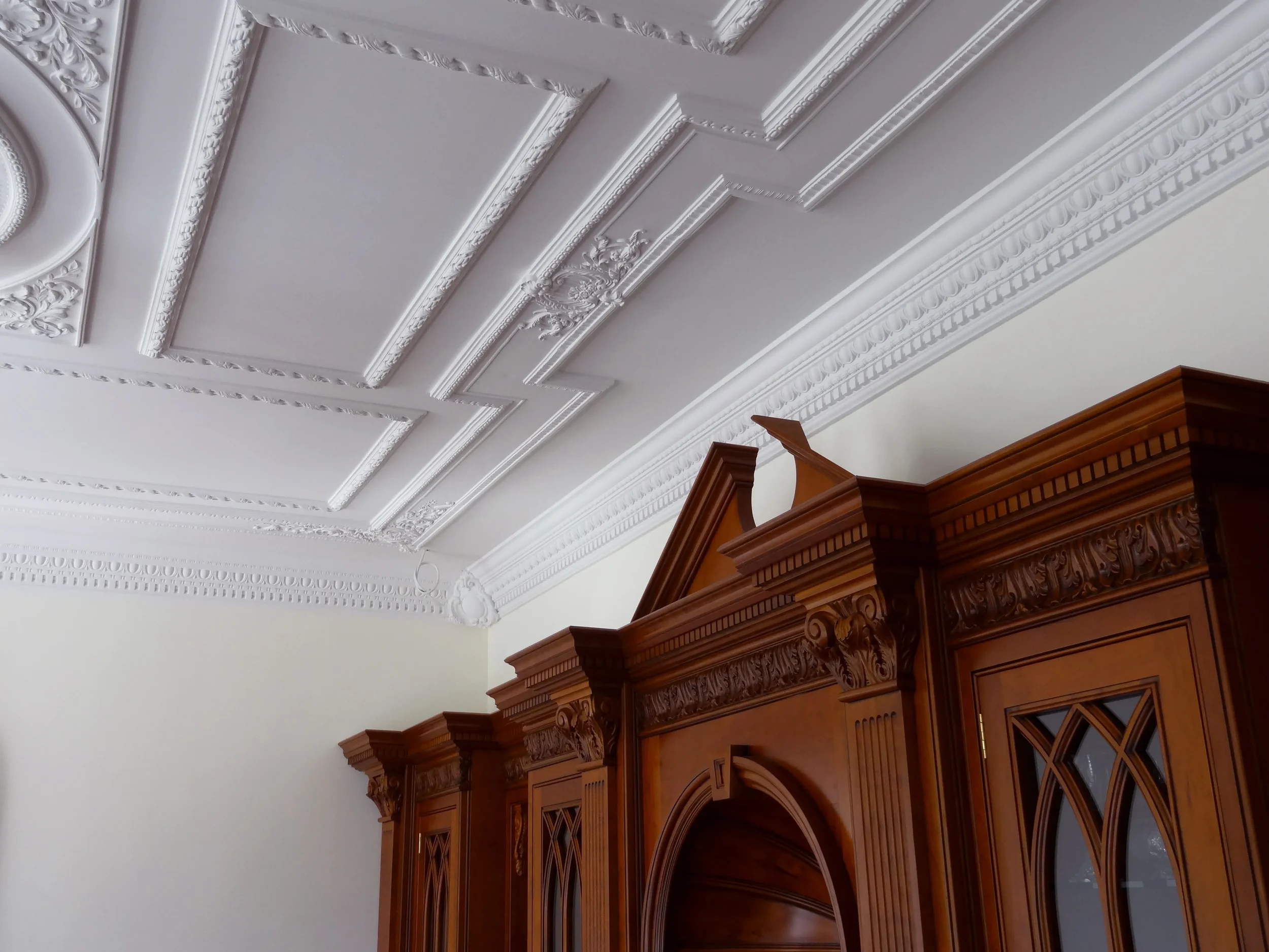Ceiling with ornate white molding and a wooden cabinet with glass doors in a room.