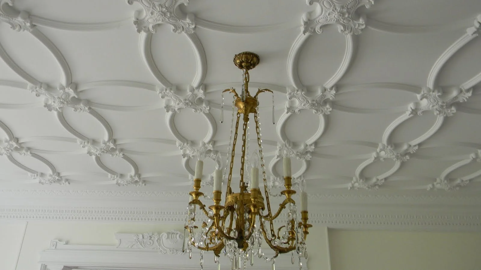 A gold chandelier with glass decorations hanging from a decorative brown ceiling mount, suspended from an ornate white ceiling with intricate molding and patterns.
