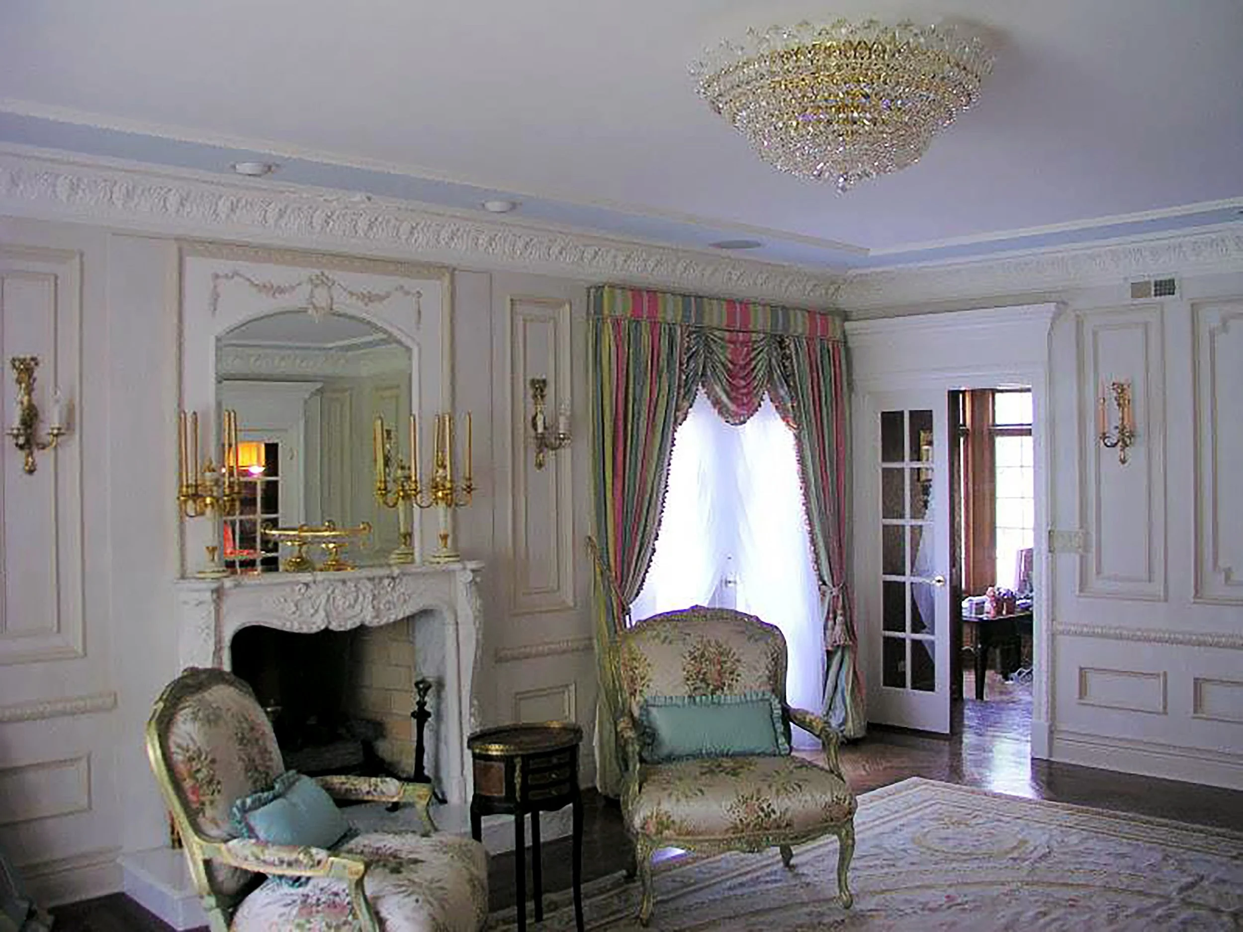 Elegant living room with ornate furnishings, floral upholstered chairs, gold accents, a fireplace with a mirror above, and a window with patterned curtains.
