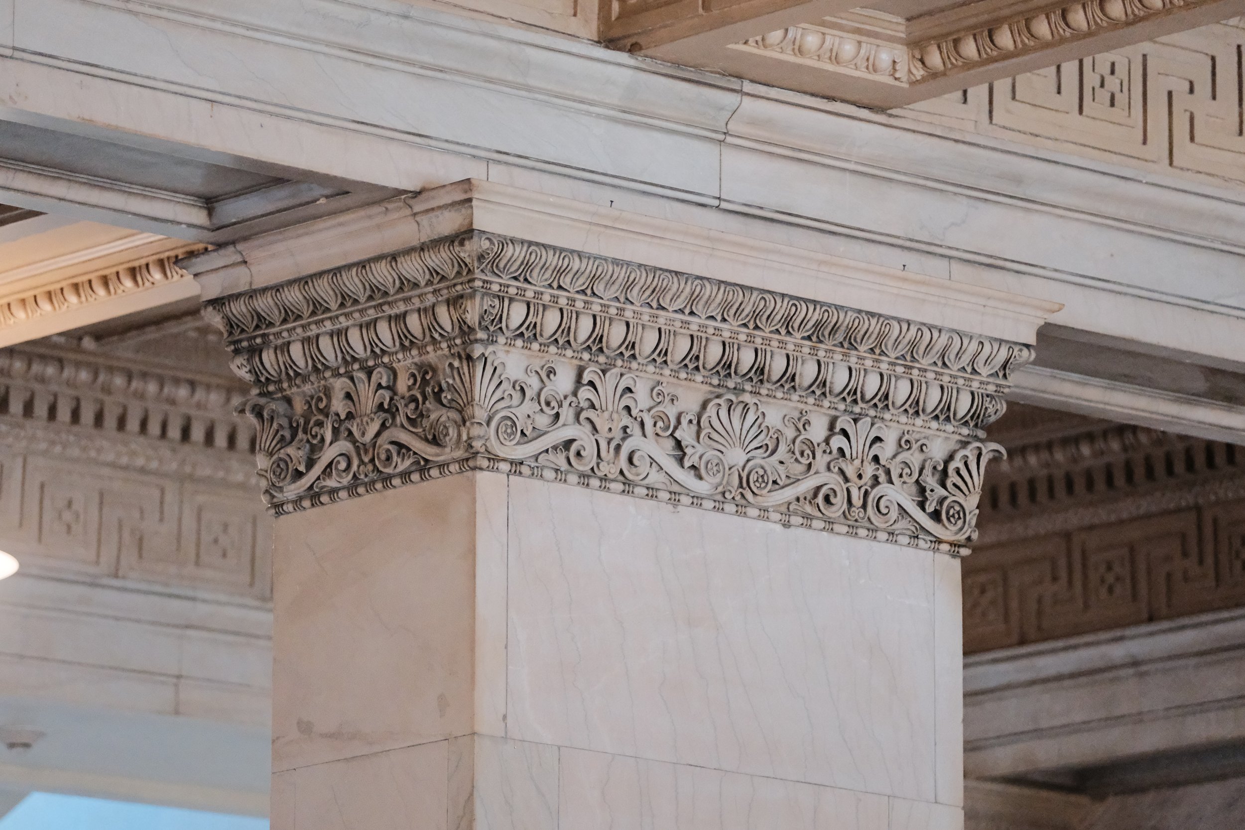 Close-up of a marble column with ornate decorative carvings at the top, including floral and scroll motifs, in classical architecture.