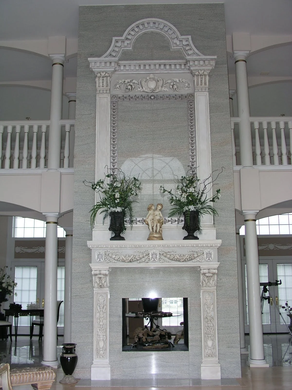 A large white marble fireplace with ornate carvings and decorative details. Two large floral arrangements in black vases sit on the mantel, with a small angel figurine between them. There is a large mirror or decorative panel above the fireplace, wit