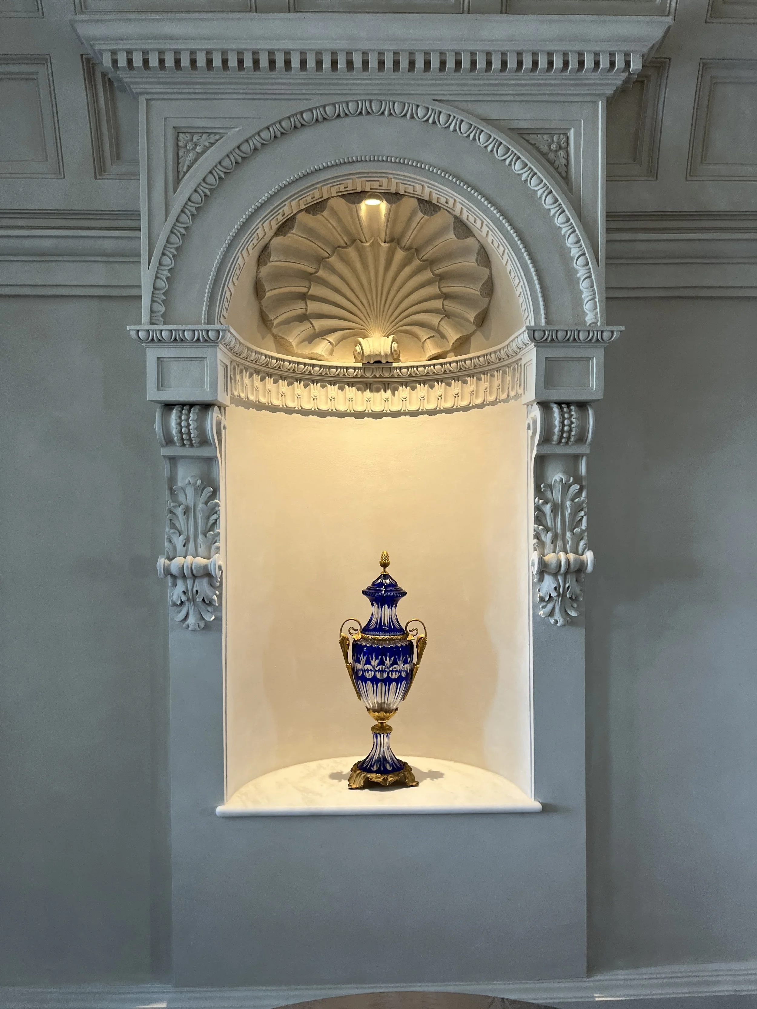 A decorative vase displayed in a lit ornate alcove in a white wall with intricate detailing.