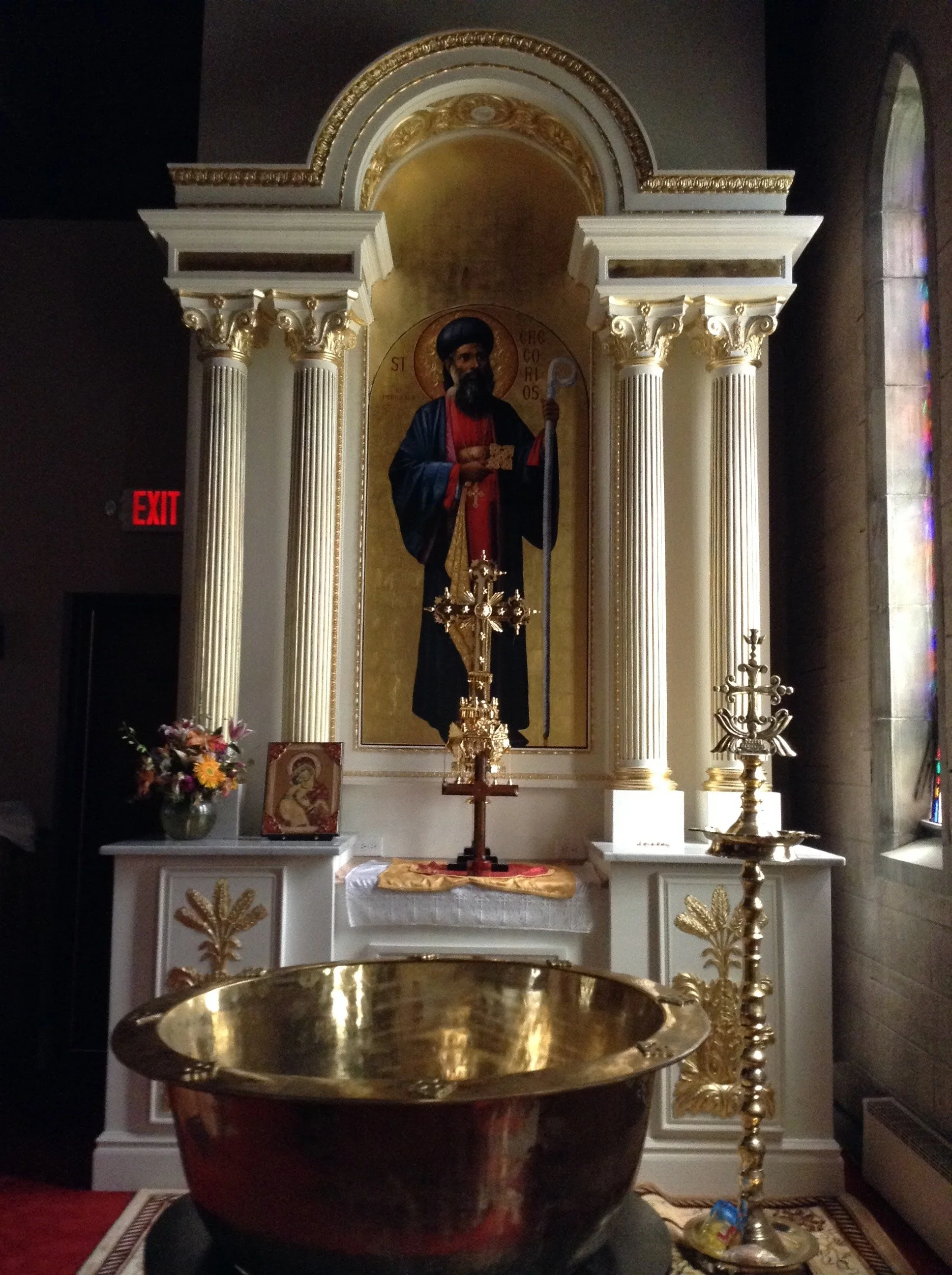 Interior of a church altar with a religious icon, candle holder, flowers, and a gold basin in the foreground.