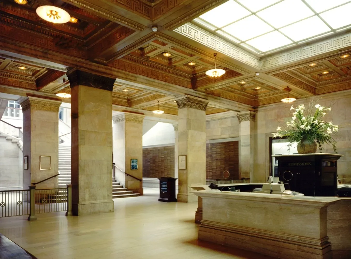 Interior of a grand building with marble columns, wooden ceiling with ornate details, staircase, vase with flowers, and a reception desk.