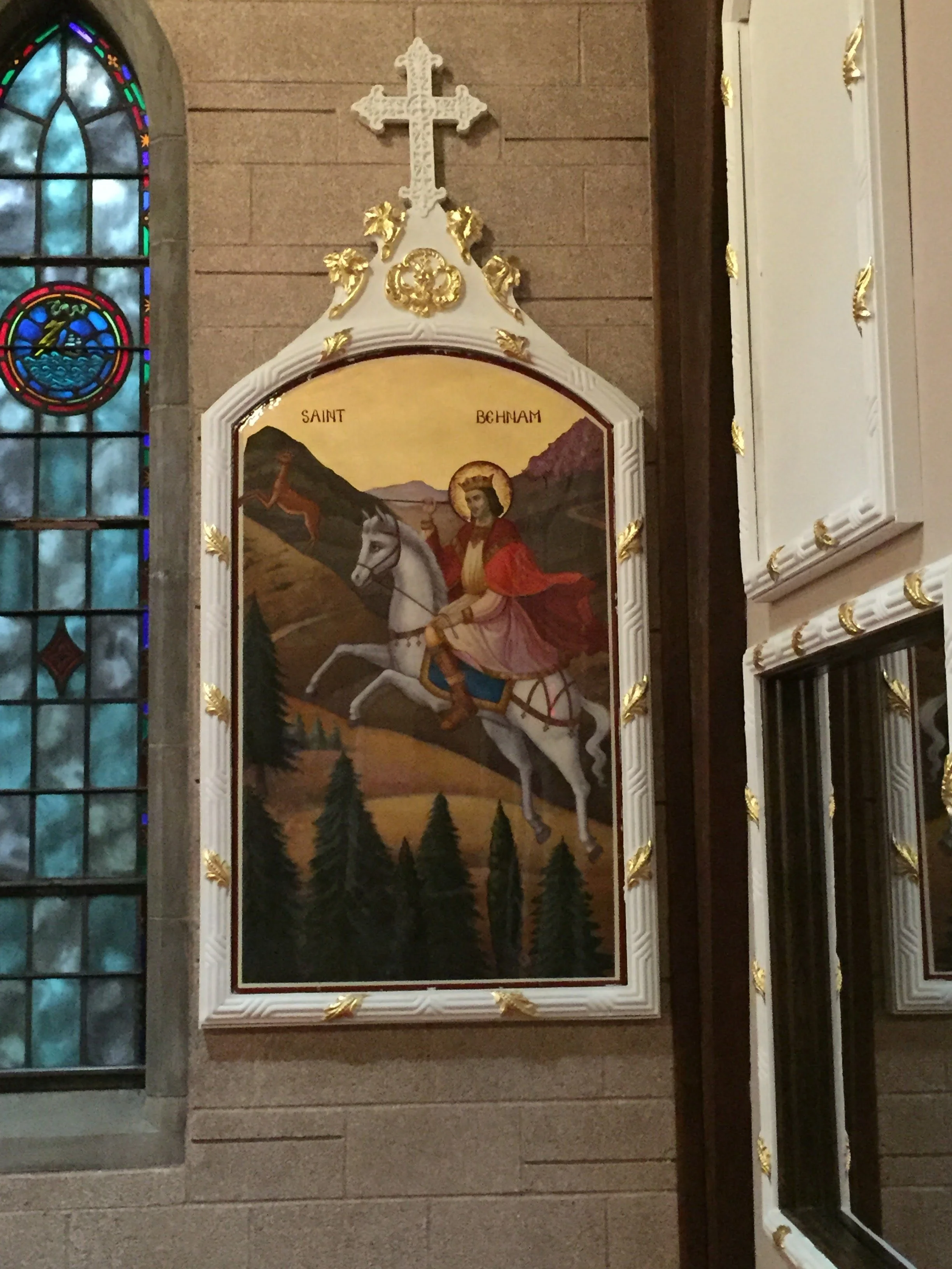 Religious painting of Saint Behnam on a white horse with a halo, set against a mountainous landscape with pine trees. The frame is ornate with gold accents, and a cross is mounted above the painting. Stained glass window is visible on the left side.