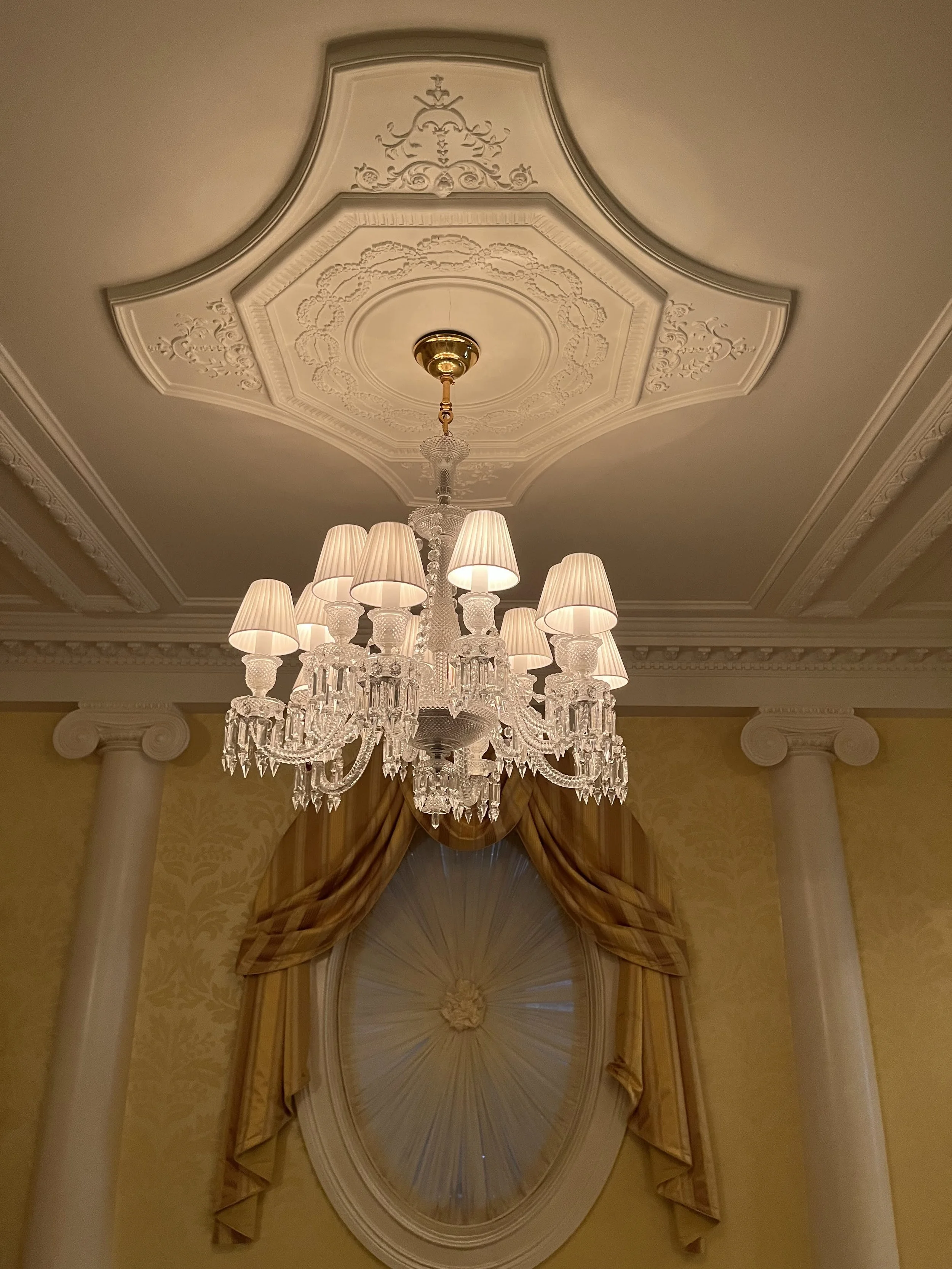 Elegant chandelier with multiple white lampshades hanging from ornate ceiling medallion in a luxurious room with draped window.