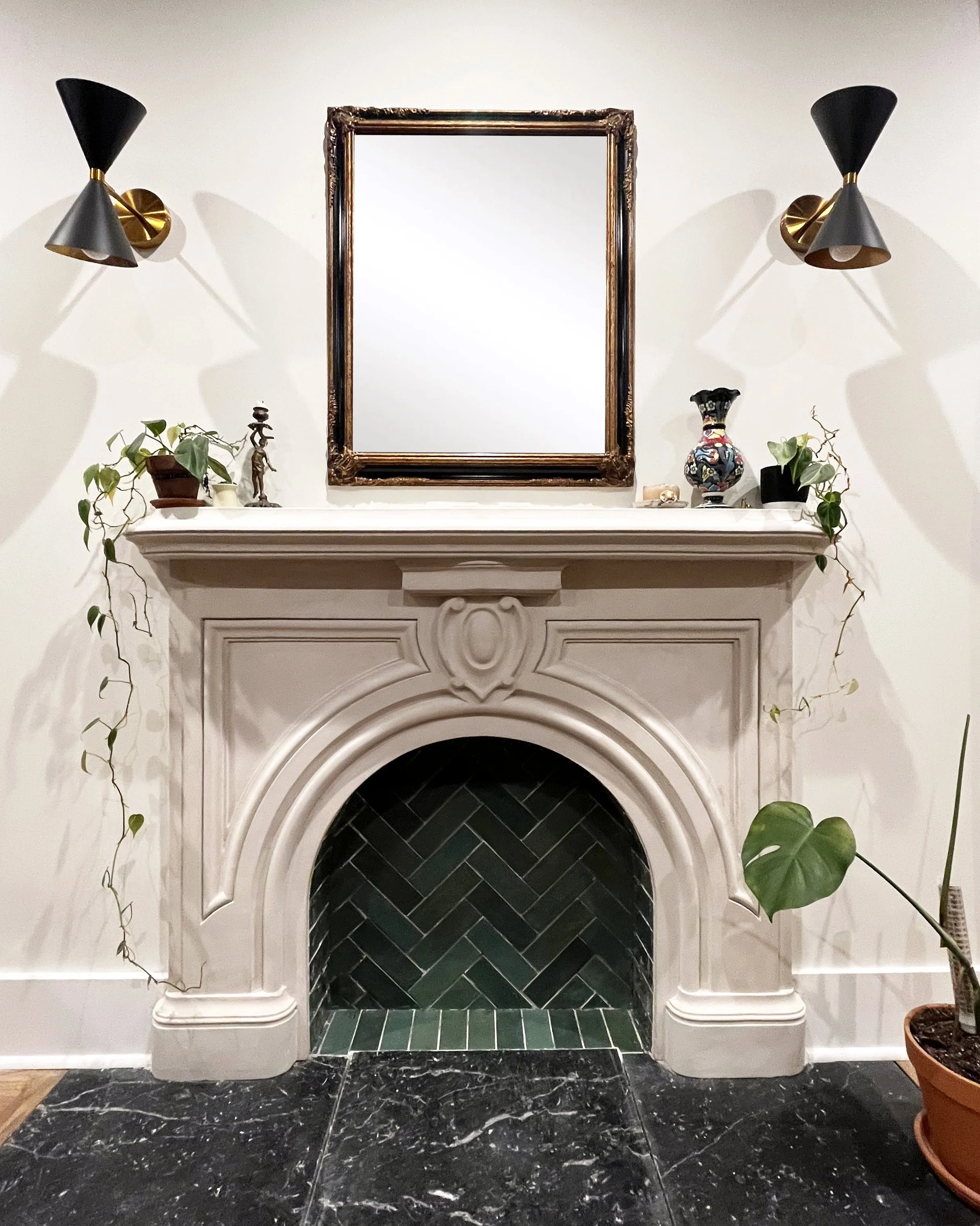 A white fireplace with black herringbone tile inside the opening, decorated with plants, a small statue, a decorative vase, and a mirror above it. Two black and gold wall sconces are mounted on each side of the mirror.