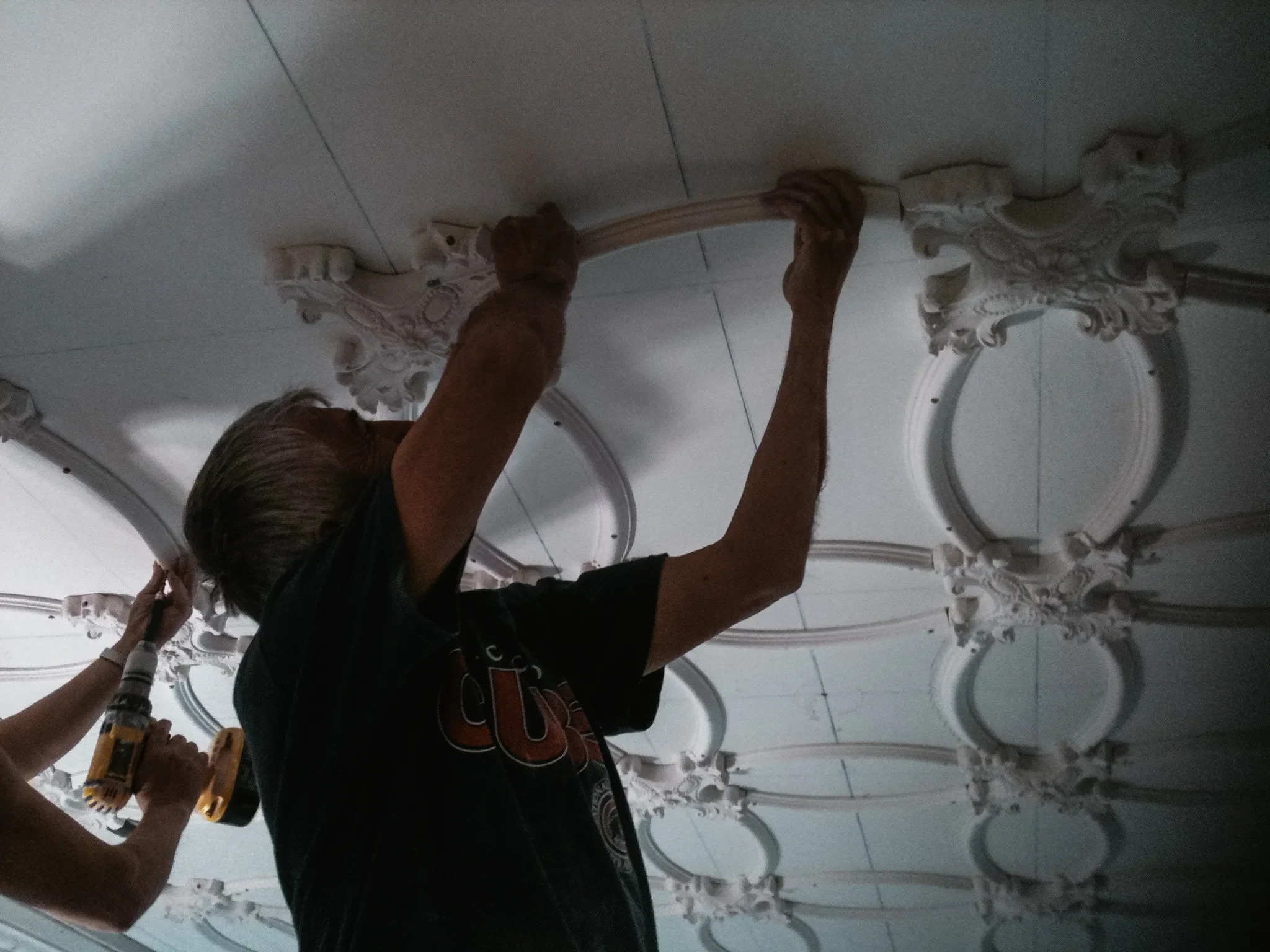 Two people are installing or repairing decorative ceiling medallions, one person is using a cordless drill while the other is holding a tool, and both are working on a ceiling with multiple ornate white medallions.