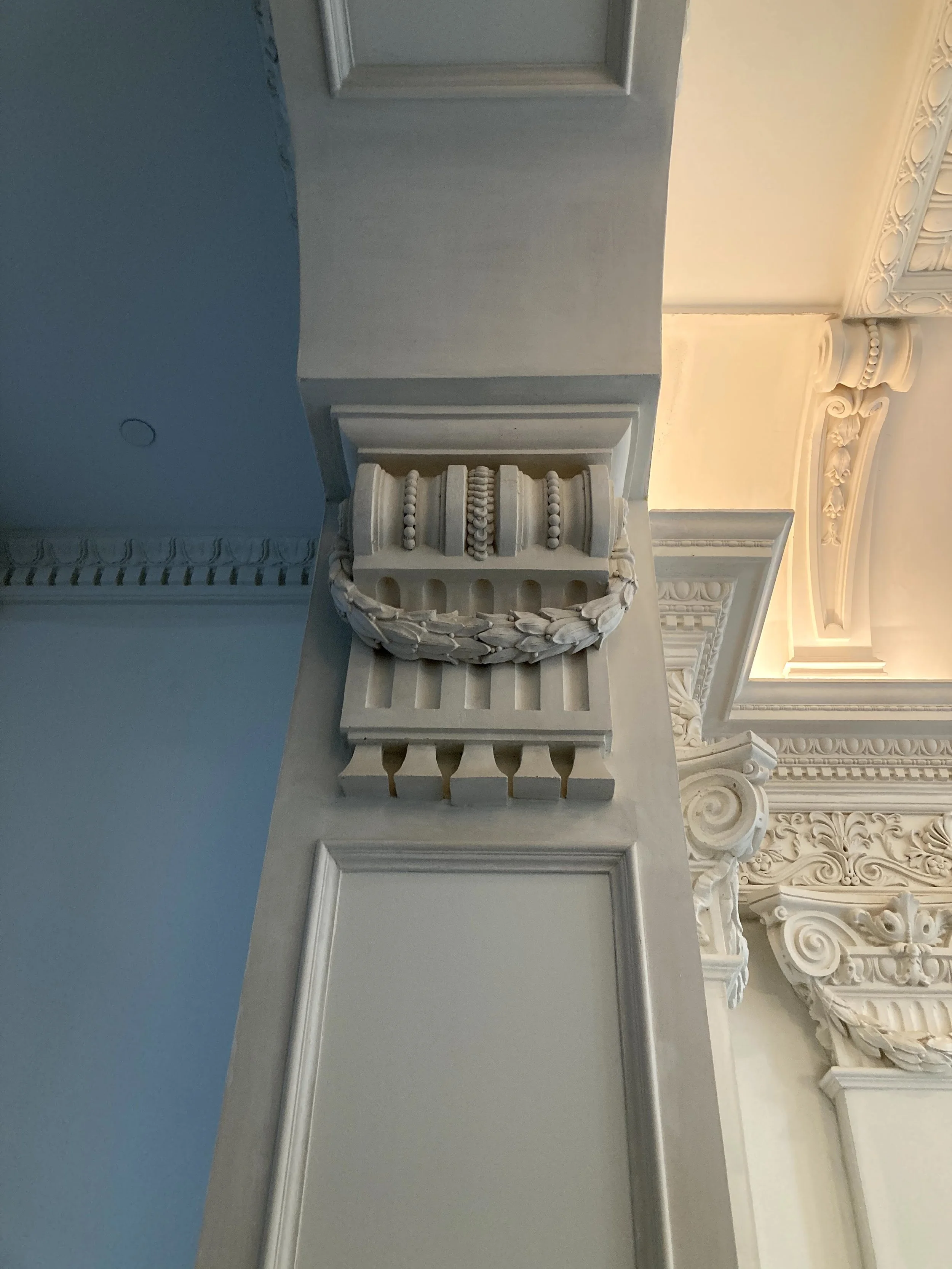 Close-up view of ornate white architectural detailing, including decorative molding and carved embellishments on a ceiling and wall, with a column featuring decorative elements.