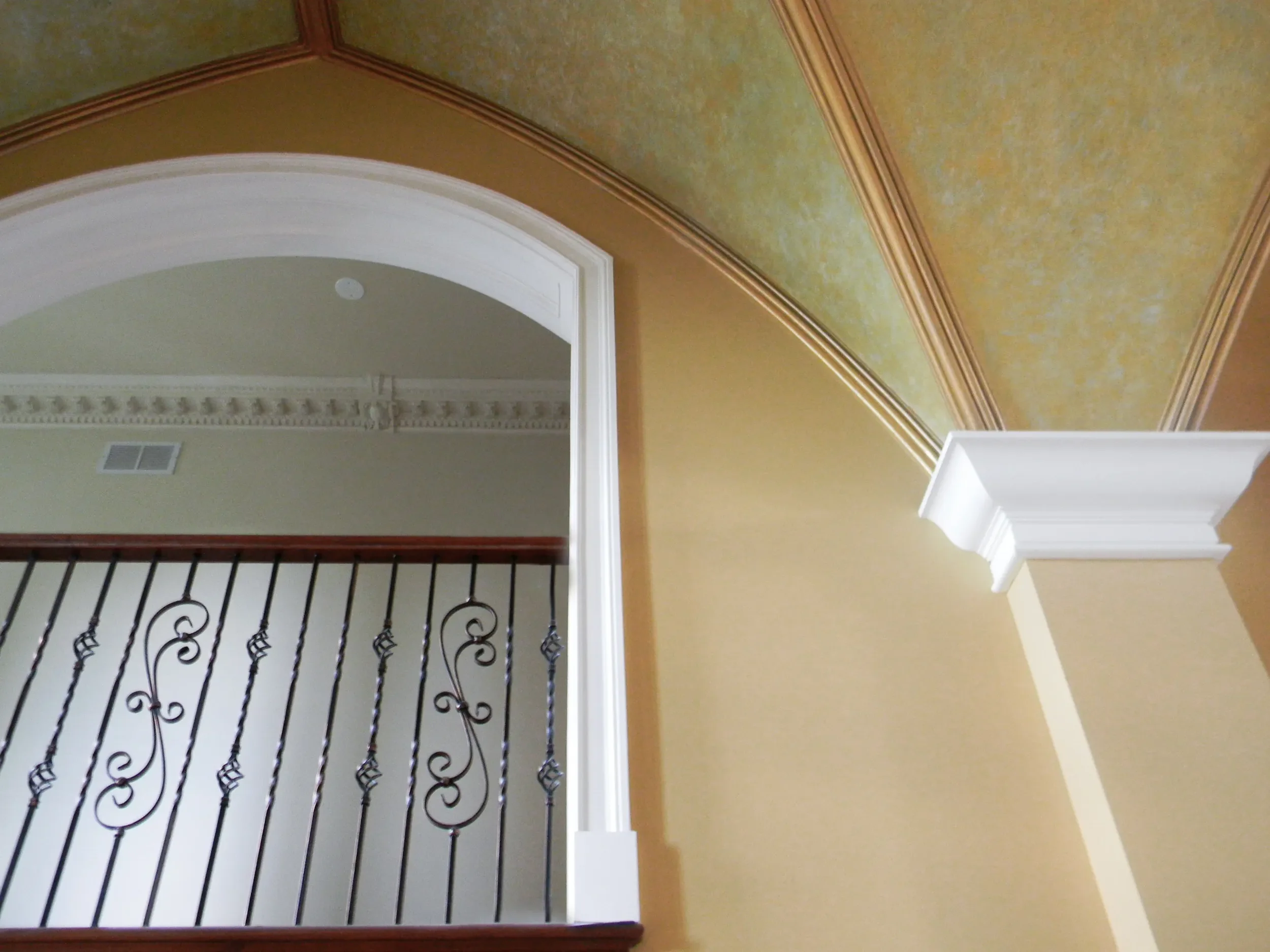 View of an interior part of a house showing an arched doorway, a decorative ceiling with gold and green tones, a white column with crown molding, and a railing with wrought iron balusters.
