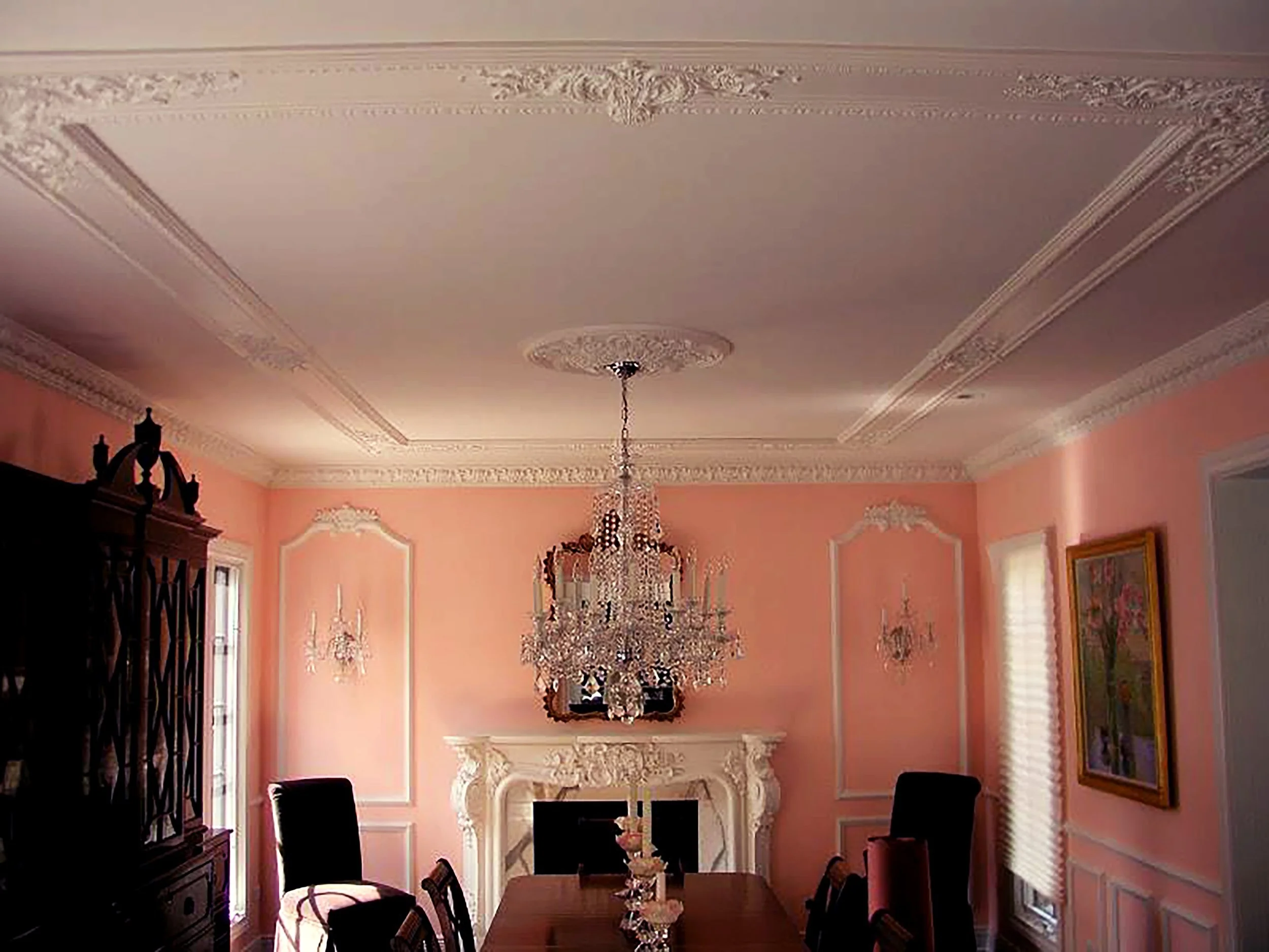 Elegant dining room with pink walls, white ornate ceiling moldings, a large crystal chandelier, and wooden dining table with black chairs. Decorative wall sconces, framed painting, white fireplace, and windows with blinds.