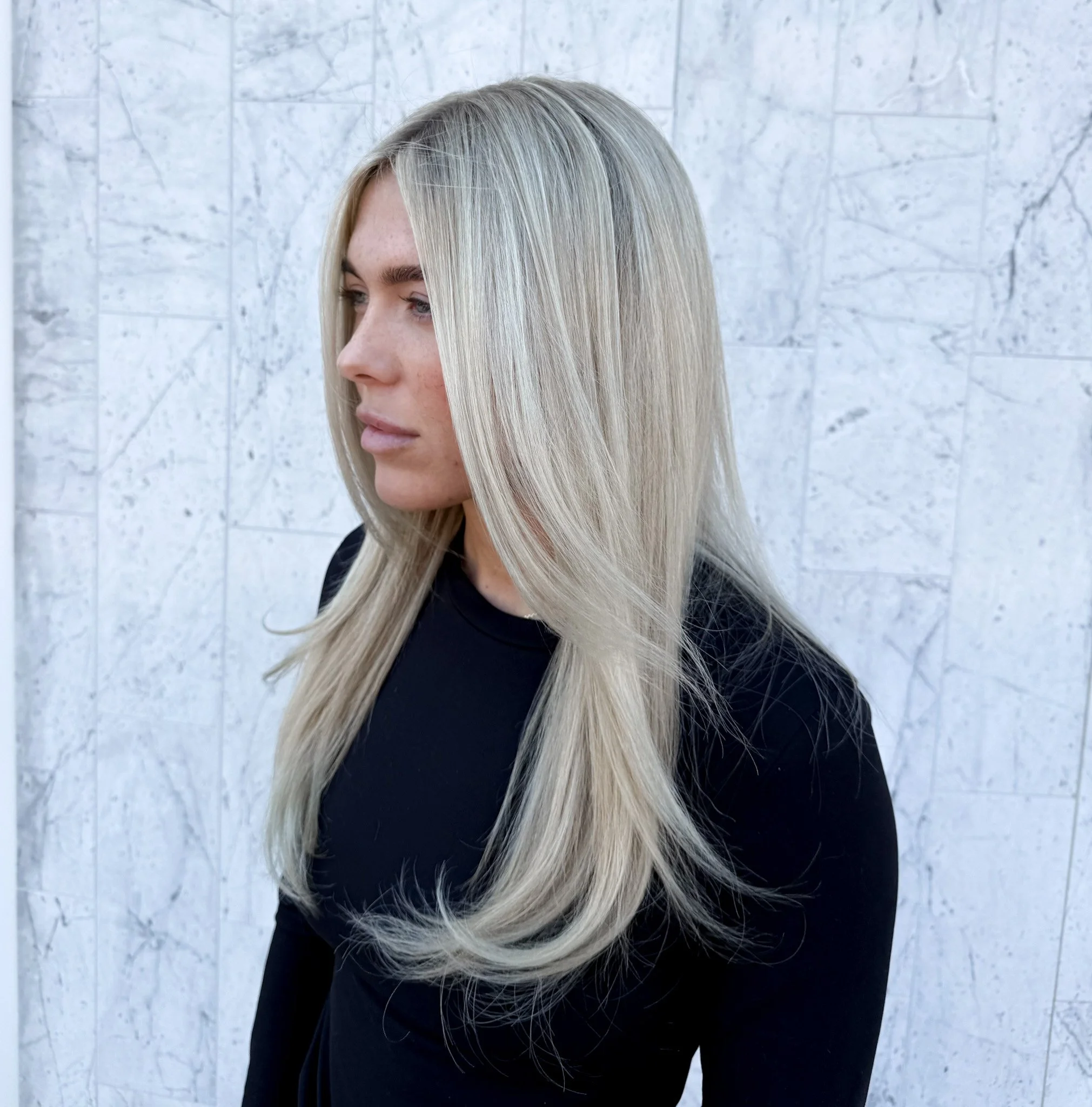 A woman with long, straight blonde hair wearing a black top, standing in front of a light-colored tiled wall.