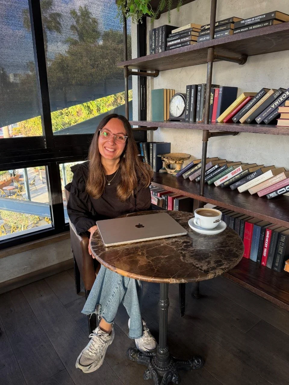 Eine junge Frau mit langen braunen Haaren, Brille und einem schwarzen Oberteil sitzt an einem runden, braunen Marmortisch in einem Café. Auf dem Tisch liegt ein Laptop und eine Tasse Kaffee. Im Hintergrund sind Bücherregale mit vielen Büchern und eine große Fensterfläche mit Blick nach draußen.