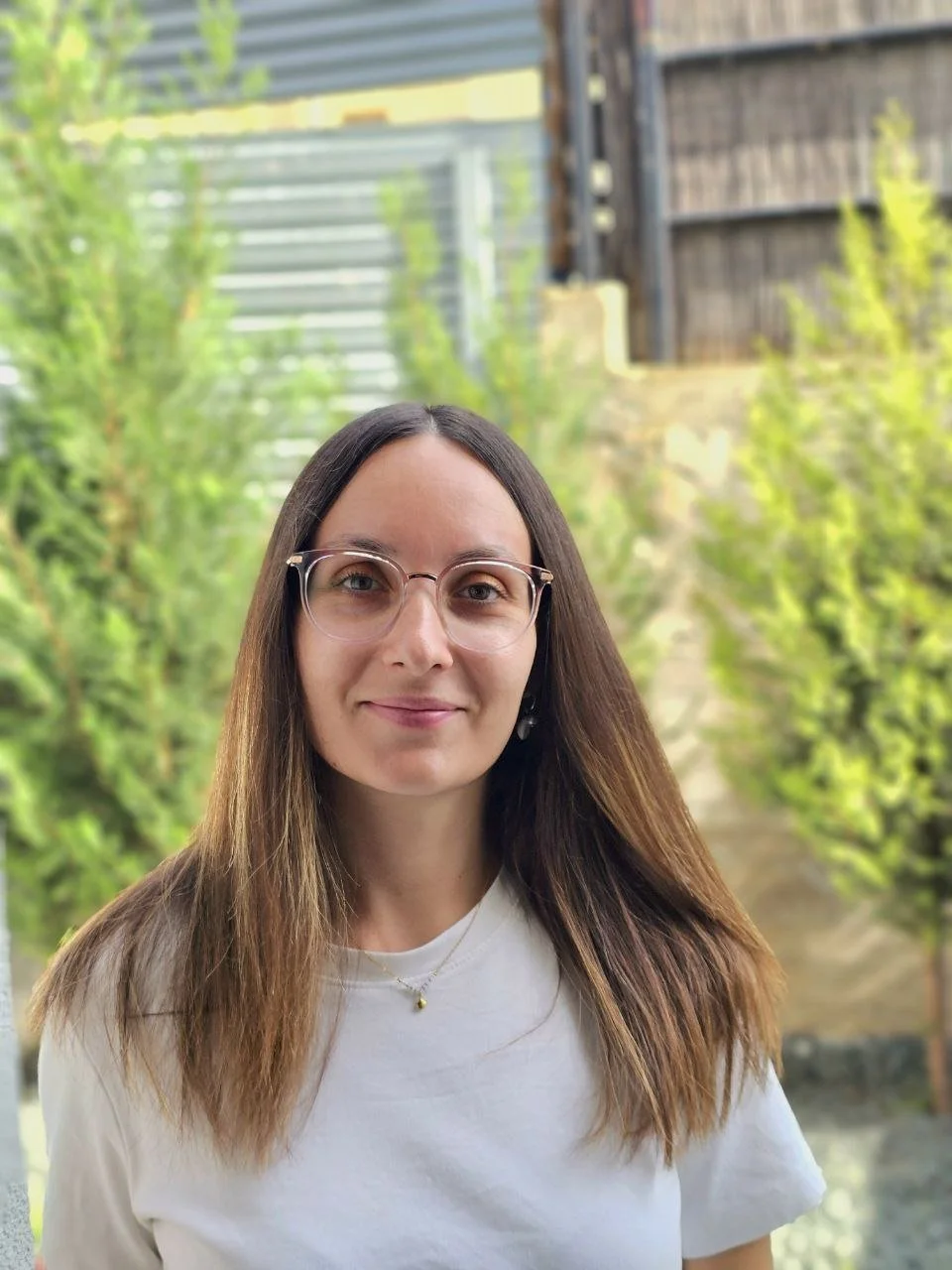 Junge Frau mit Brille und langen braunen Haaren, lächelnd, im Garten mit grünen Pflanzen, Holz und Metallzaun im Hintergrund.