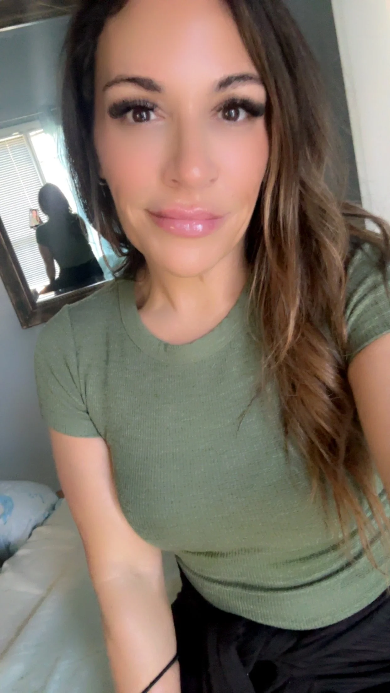 A woman with long wavy brown hair, wearing a green t-shirt, smiling at the camera in a well-lit room. A mirror and a person taking the photo are visible in the background.