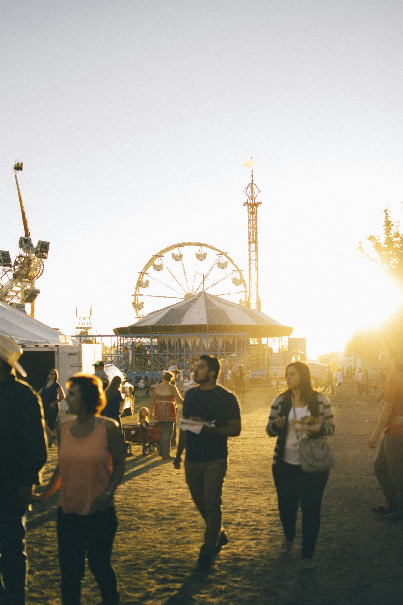 People walking in a carnival with a Ferris wheel and amusement rides in the background during sunset.