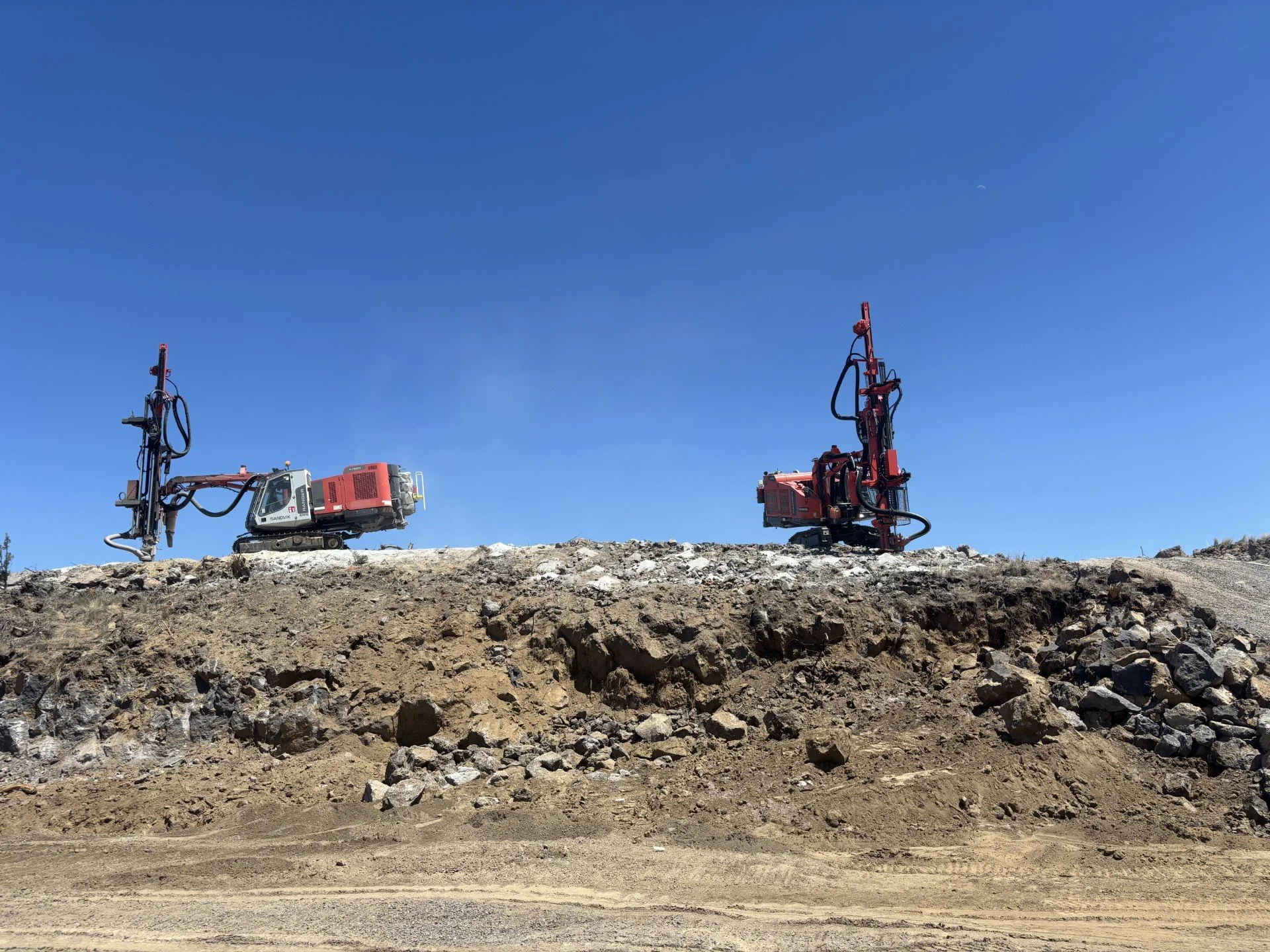 Two large drilling machines on rough terrain, with a clear blue sky in the background.