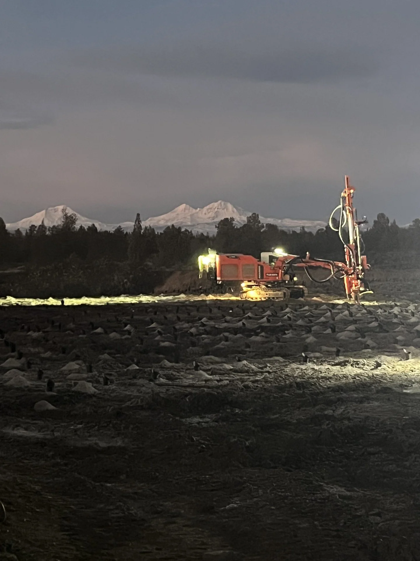 A large red drilling machine working on a dark, rocky terrain at dusk or dawn with snow-capped mountains in the background.