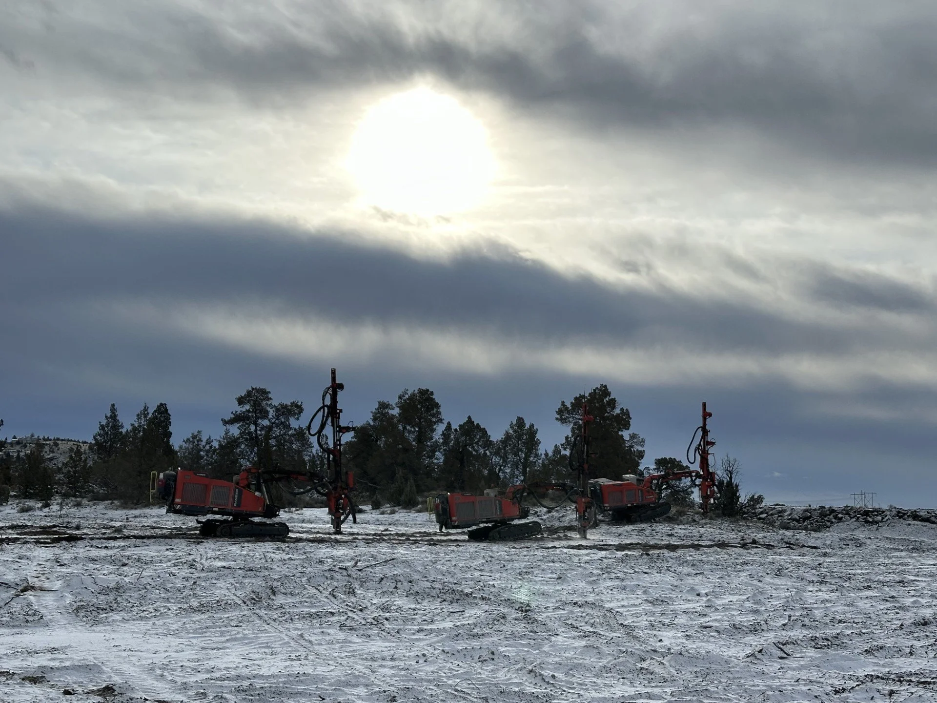 Three red tracked drilling machines in a snowy landscape with trees and a cloudy sky with the sun shining through