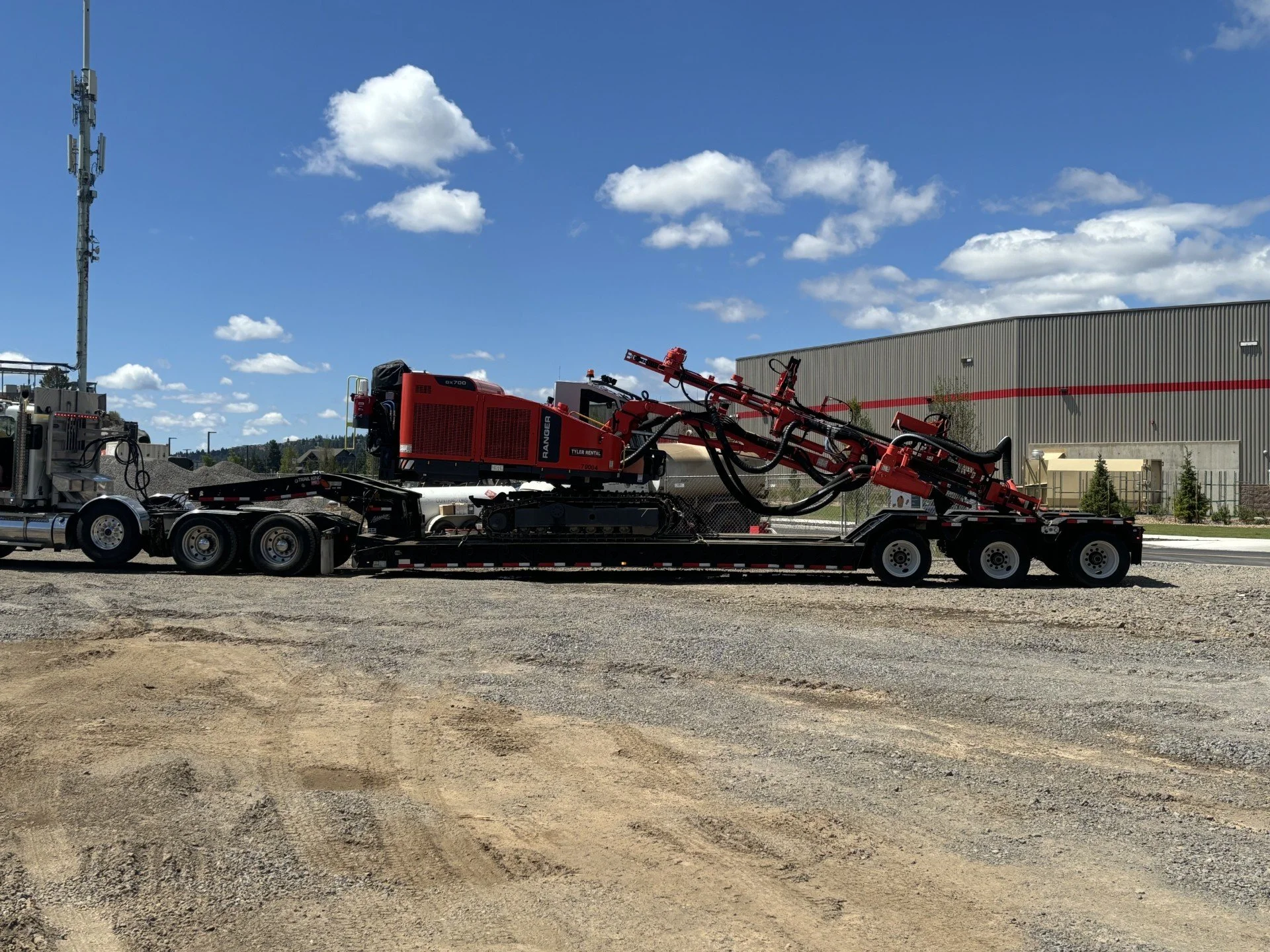 A large red and black industrial machine on a flatbed trailer parked on a gravel lot with a warehouse in the background and a blue sky with scattered clouds.