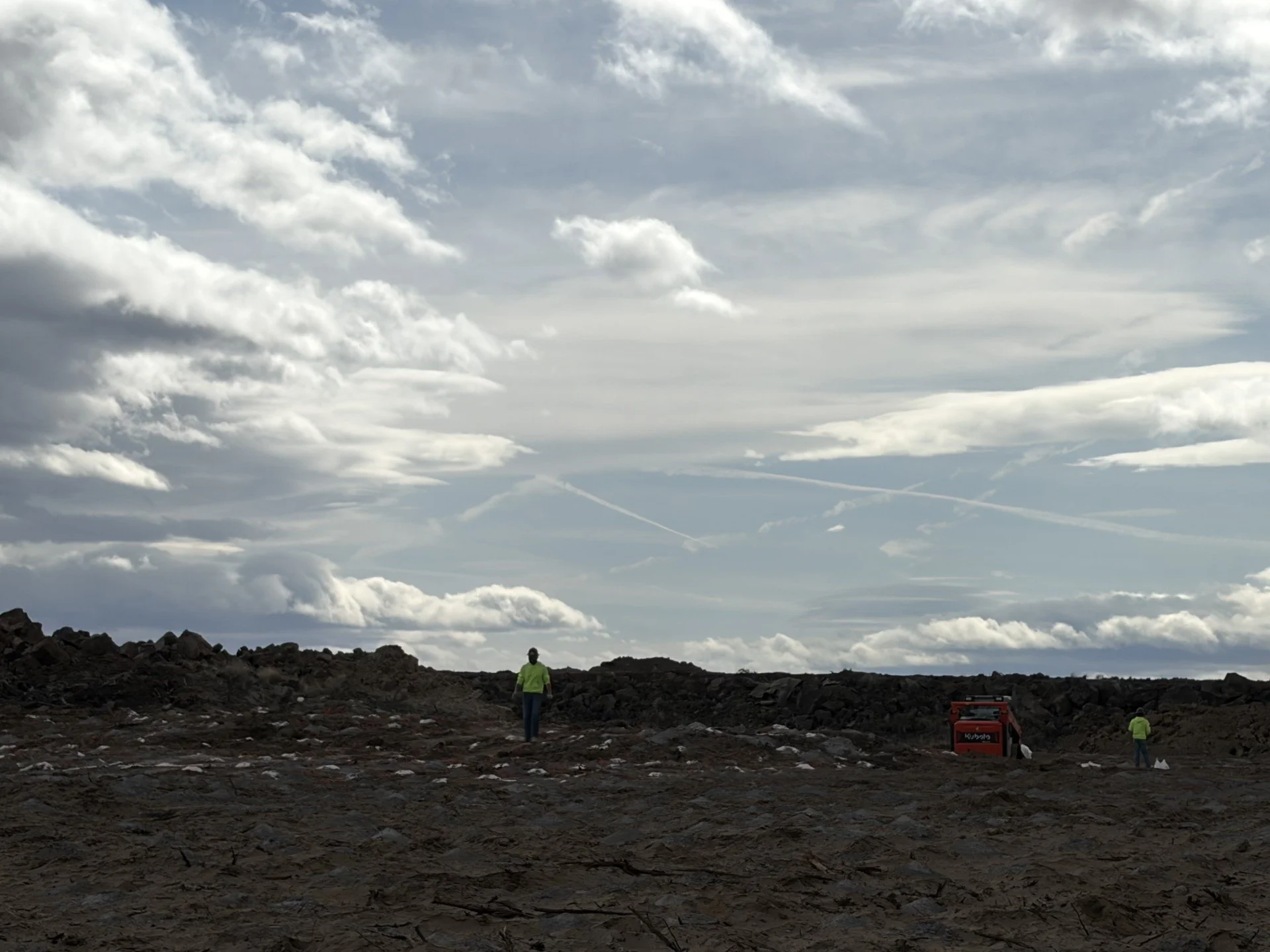 Two workers wearing safety vests on a dirt landscape under a cloudy sky with airplane contrails, and a red container or machine in the background.