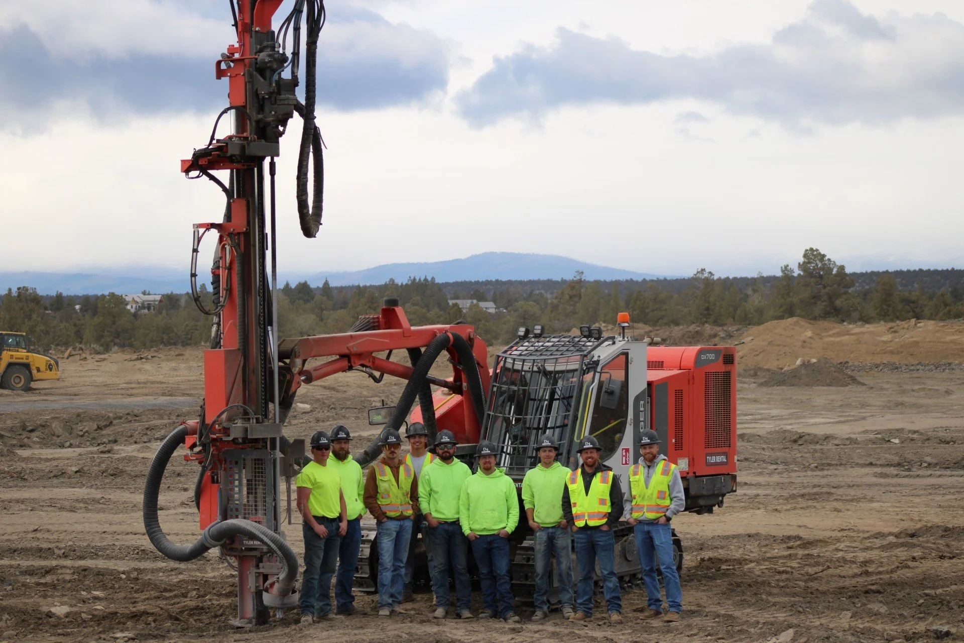 Group of nine construction workers standing in front of a large red drilling machine on a construction site with an overcast sky.