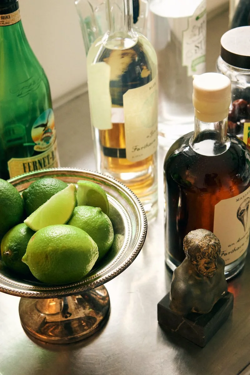 A collection of liquor bottles, fresh limes in a silver bowl, and a small bust statue on a table.