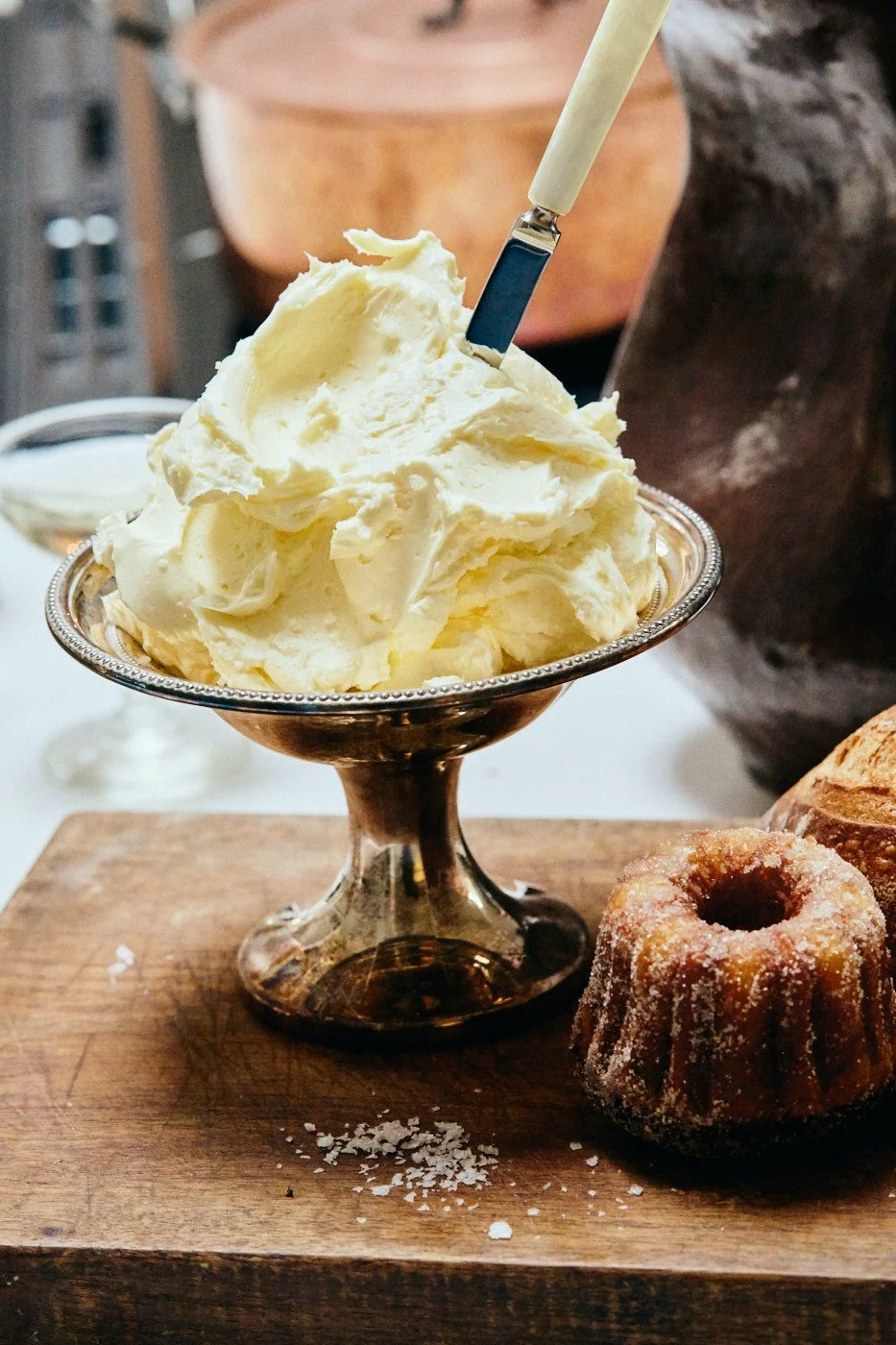 A silver dessert dish filled with vanilla ice cream, with a spoon inserted, placed on a wooden board alongside a cinnamon sugar donut and a croissant.