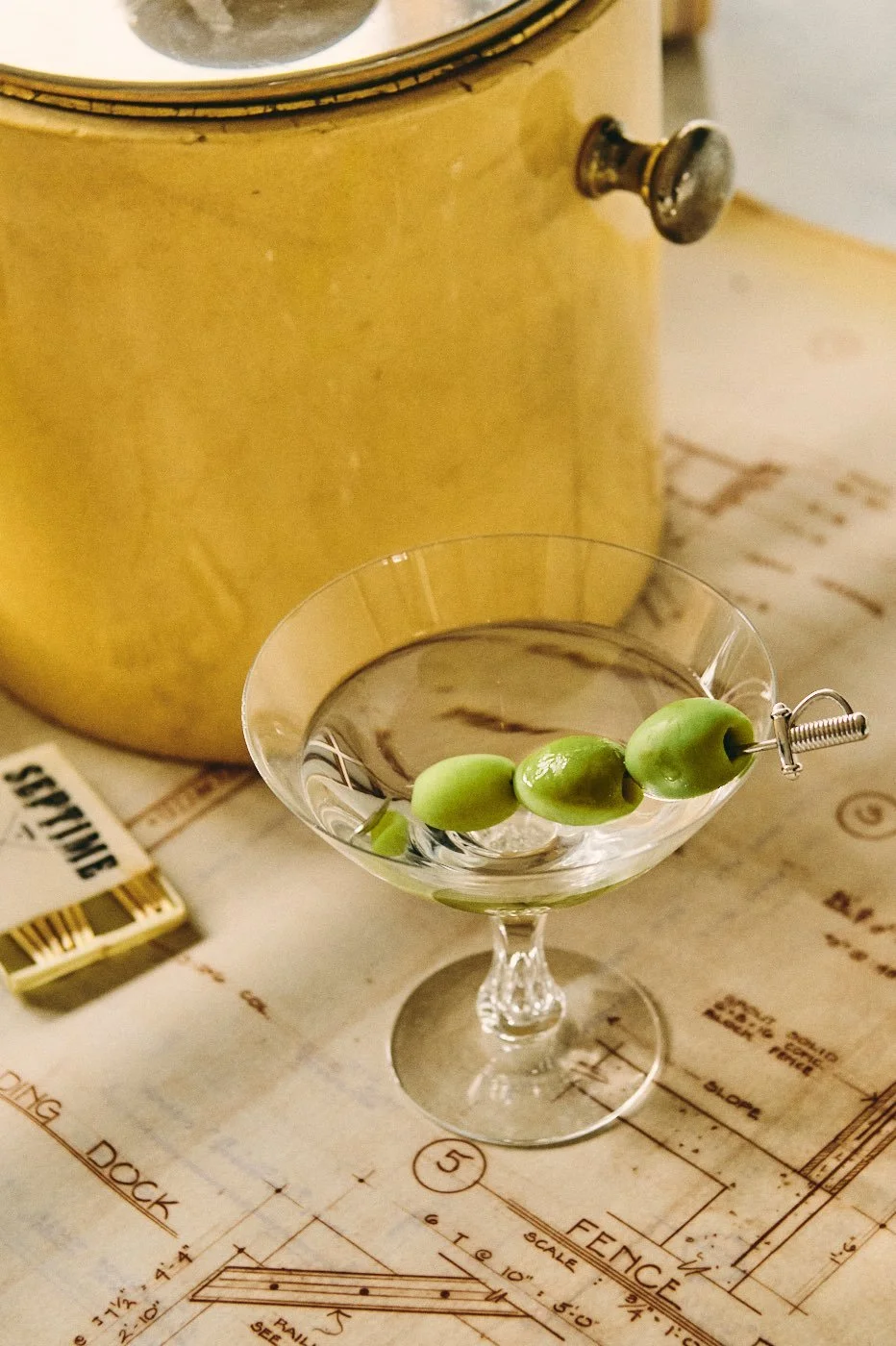A bowl of green olives with a toothpick on a glass cocktail dish, placed on a table with architectural blueprints. A yellow appliance is partially visible in the background.