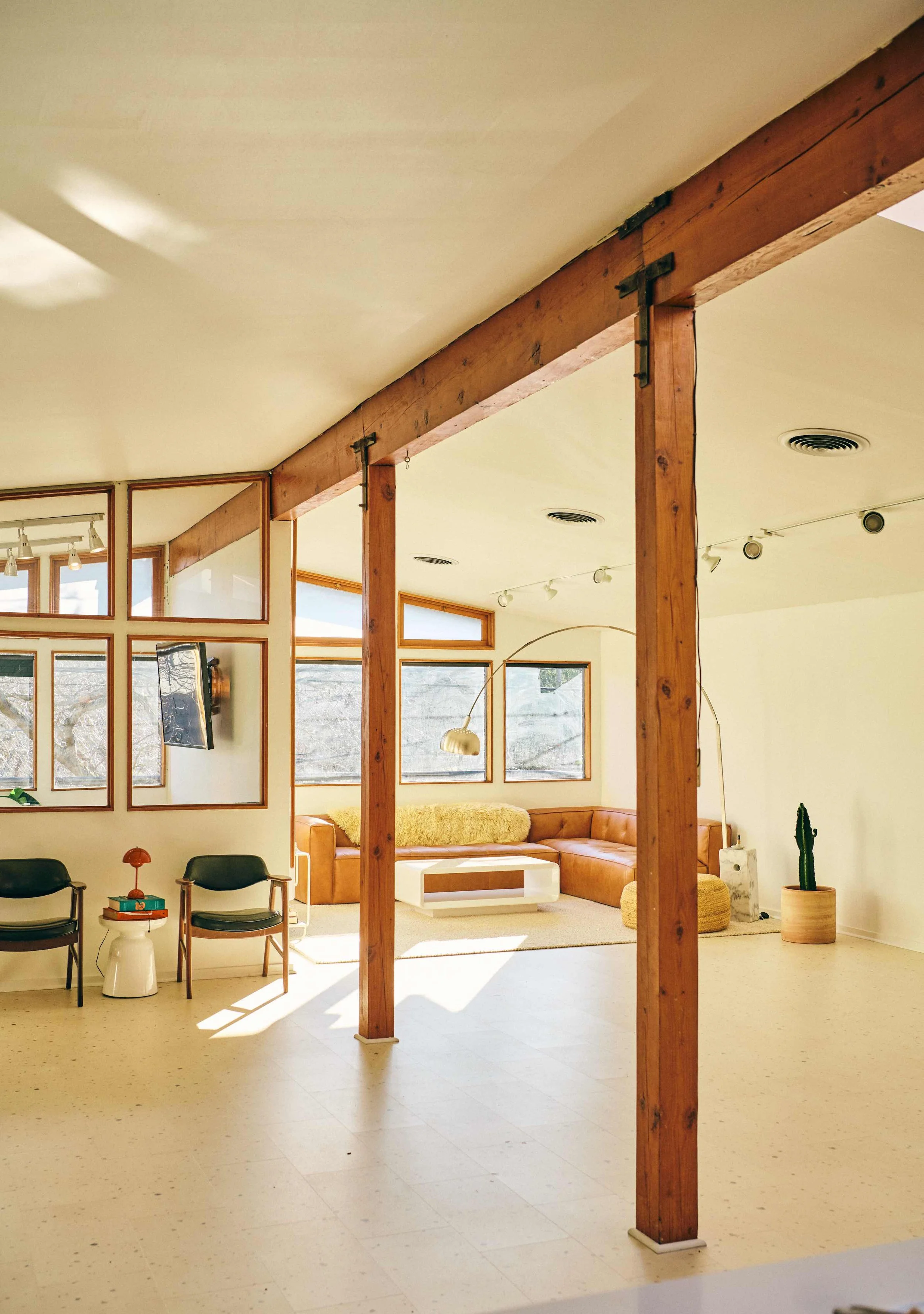 Bright, modern living space with large windows, wooden beams, a sectional sofa, armchairs, small side tables, and decorative elements like a cactus and a floor lamp.