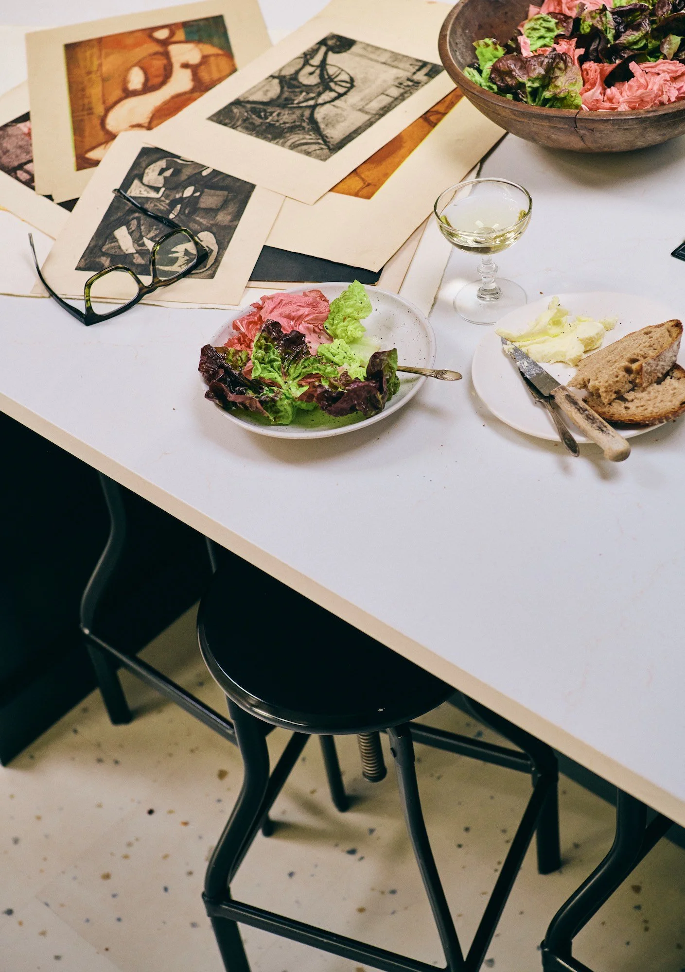 A white table with a pair of black glasses, torn bread with butter, a small glass of white wine, a bowl of mixed salad greens, and some scattered papers and artwork.