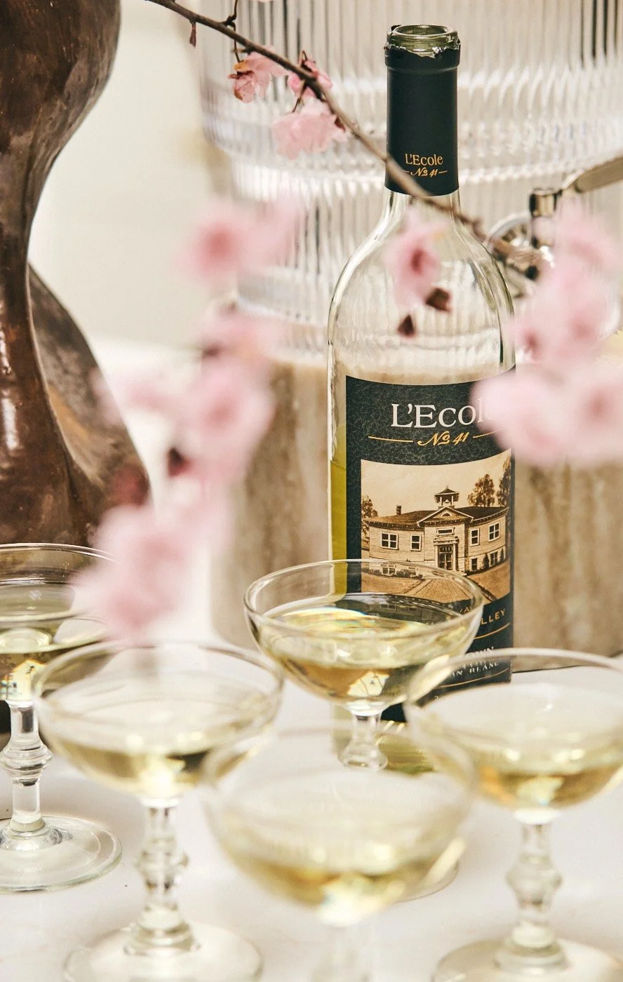 A partially empty bottle of L'Ecole No. 41 wine surrounded by several filled wine glasses, with pink cherry blossoms and decorative objects in the background.