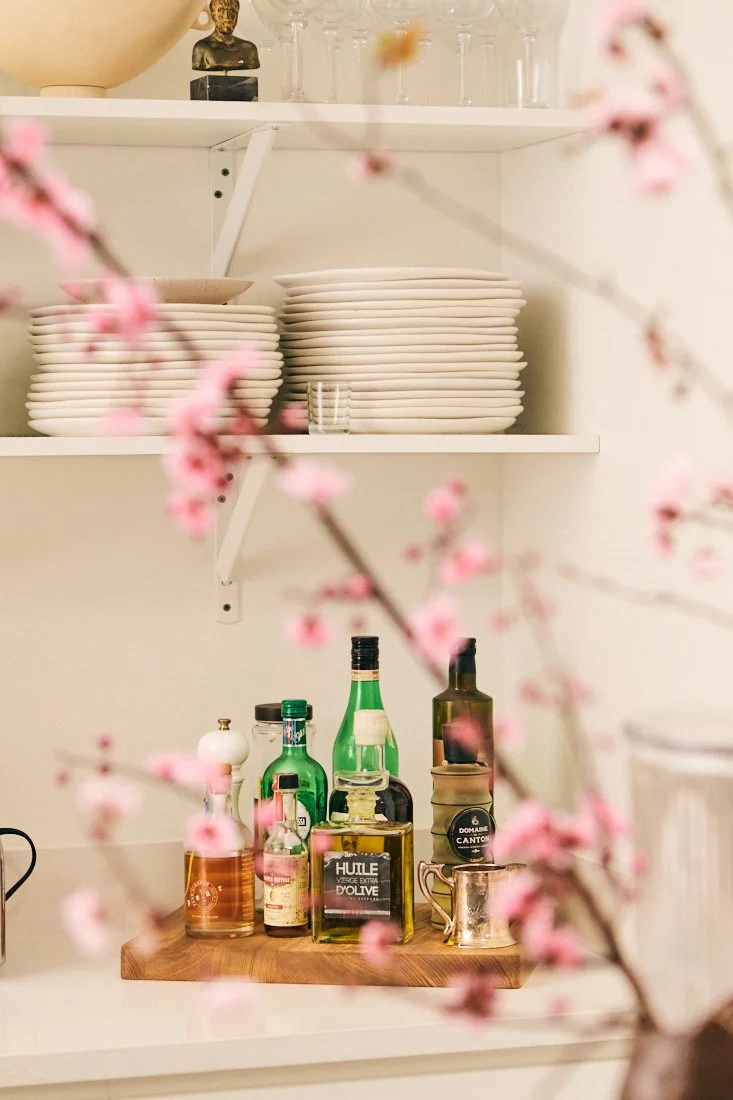 Shelves with white plates and glassware, bottles of cooking oils and condiments, and a small cup, with pink flowering branches in the foreground.