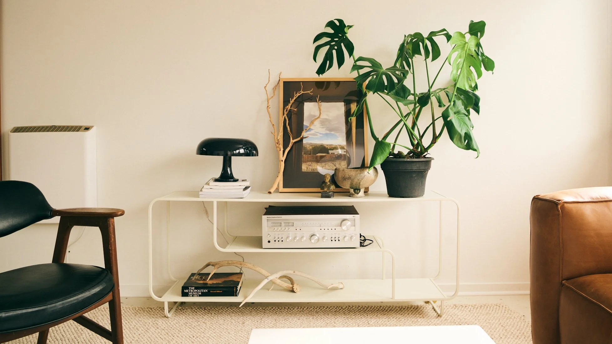 Interior living room with minimalist white console table holding framed art, plants, sculpture, and vintage stereo, with black leather chair on the left and brown leather sofa on the right.
