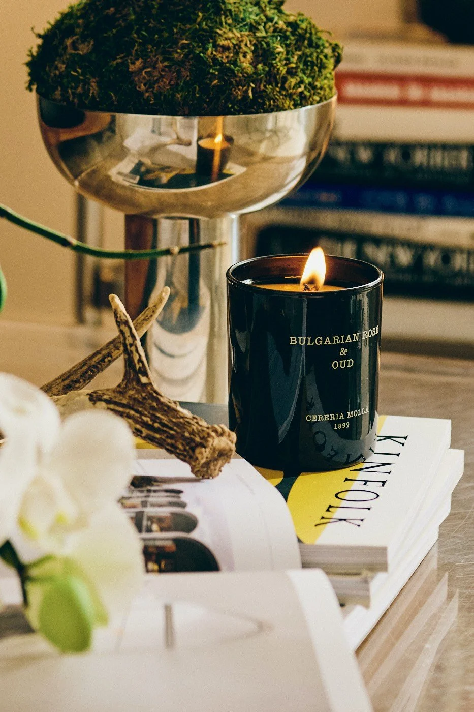 A lit black candle labeled 'Bulgarian Rose & Oud,' placed on a stack of magazines, is part of a decor arrangement that includes a metallic bowl with green moss, a small decorative branch, white flowers, and a bookshelf in the background.