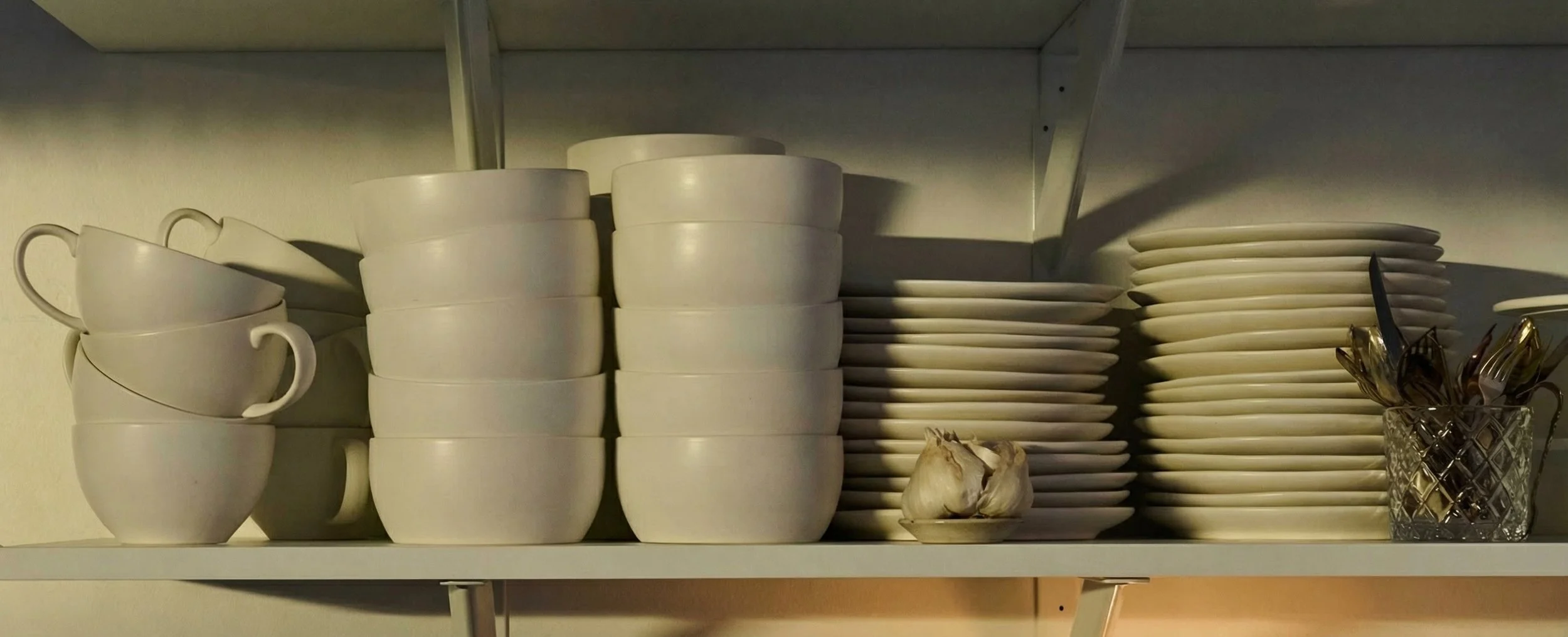 Stacked white ceramic cups, plates, and bowls, with a small garlic bulb and a glass container filled with utensils on a kitchen shelf.