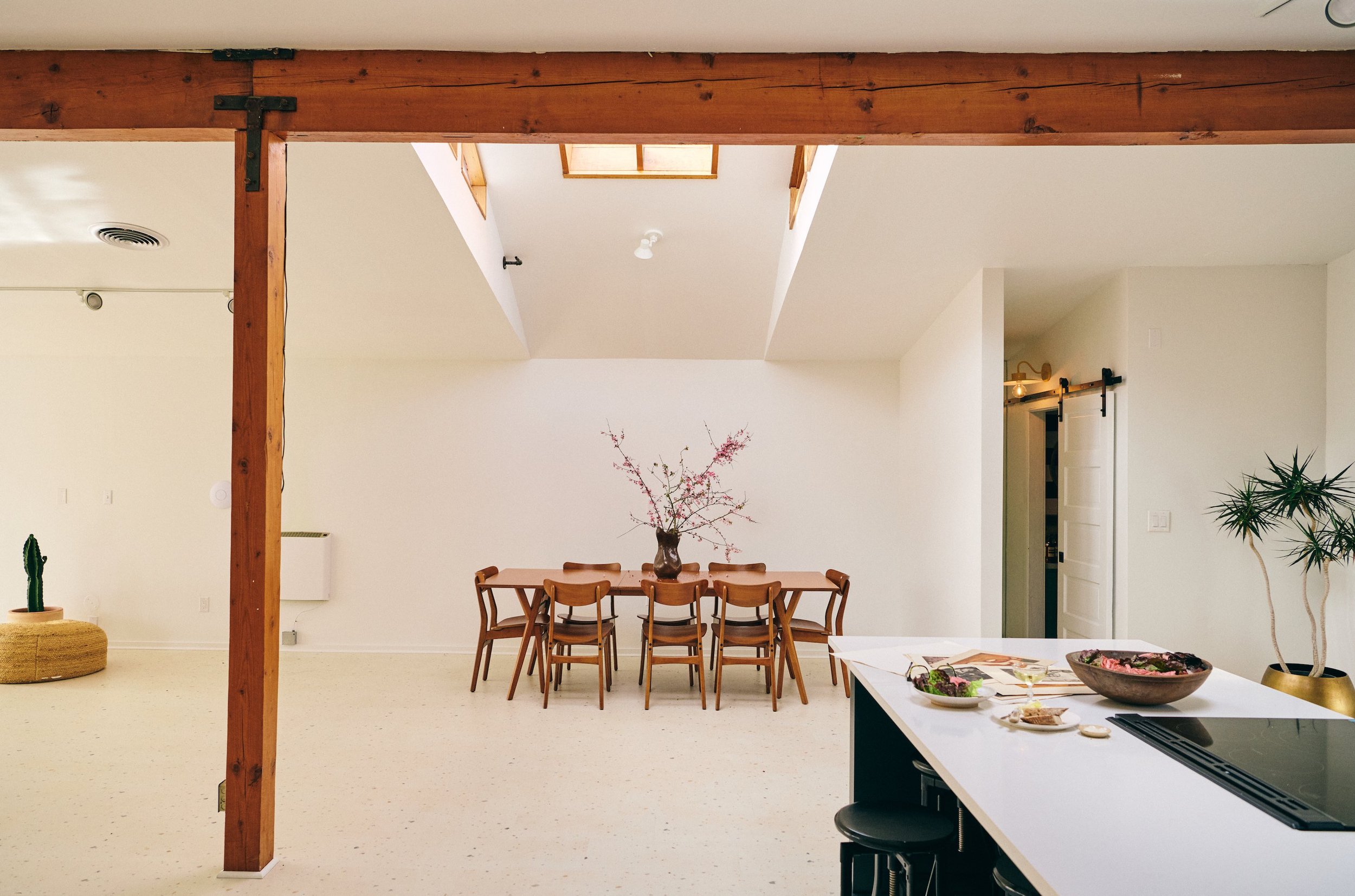 Bright and minimalistic dining area with a large wooden table, surrounded by eight wooden chairs. A vase with pink blossoms is on the table. To the right, part of a kitchen island with bowls and plates, and a plant in a gold pot. The space features w