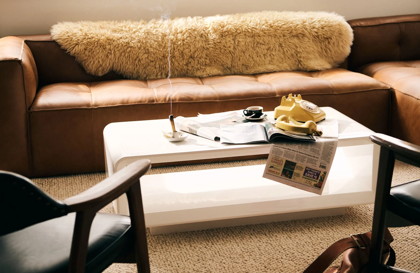 Living room with a brown leather sofa, cream throw pillow, white coffee table, vintage beige rotary phone, opened magazines, black coffee cup, and a small white dish with a lit incense stick.