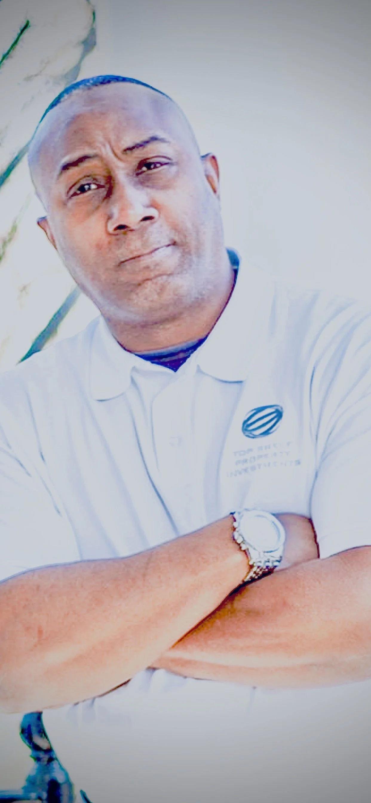 A man with crossed arms wearing a white polo shirt and a watch, standing outdoors with a neutral expression.