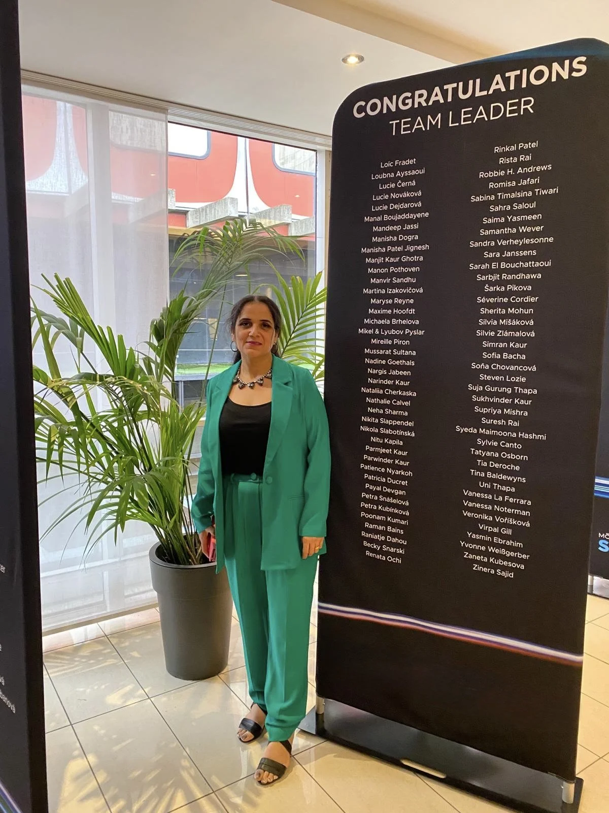 A woman in a teal suit and black top standing indoors next to a large congratulatory team leader signboard with a list of names, and a large potted plant, near a glass window with a view of a building outside.