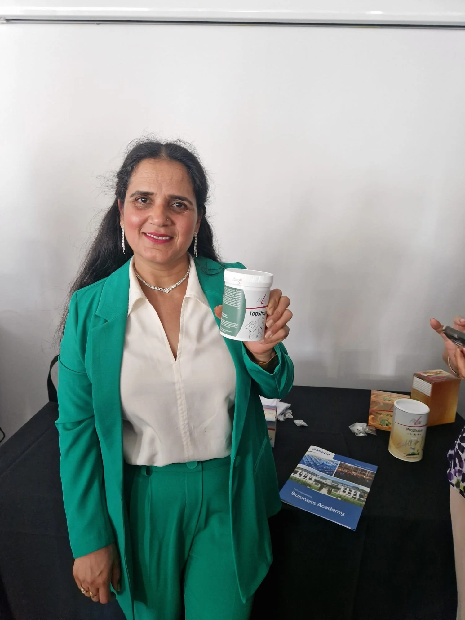 A woman with dark wavy hair, wearing a green blazer and matching pants, holding a container of a supplement or medication, smiling, standing in front of a table with promotional materials.