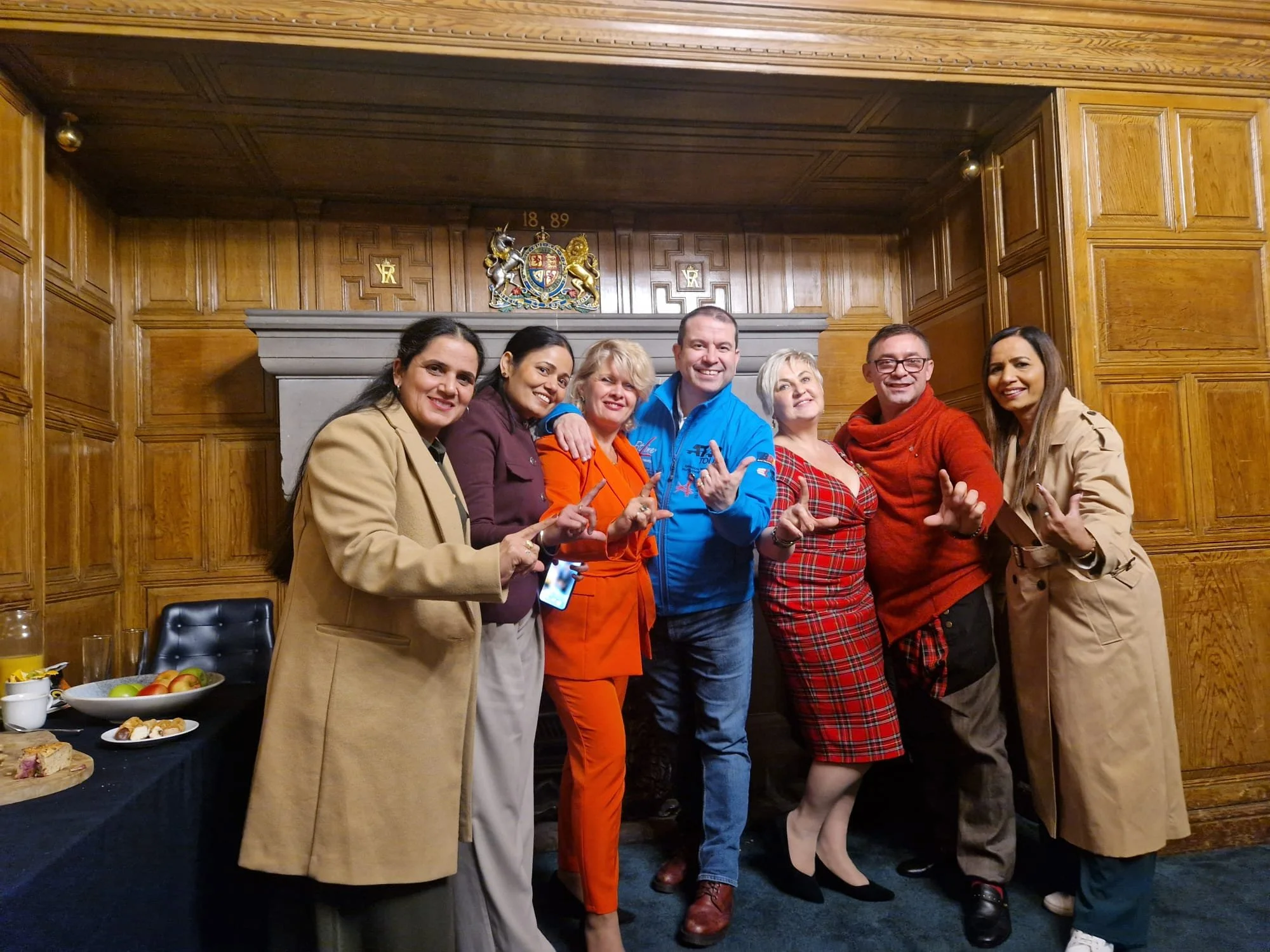 Group of seven people standing in front of a wooden wall, celebrating and smiling, with a table of food on the left.