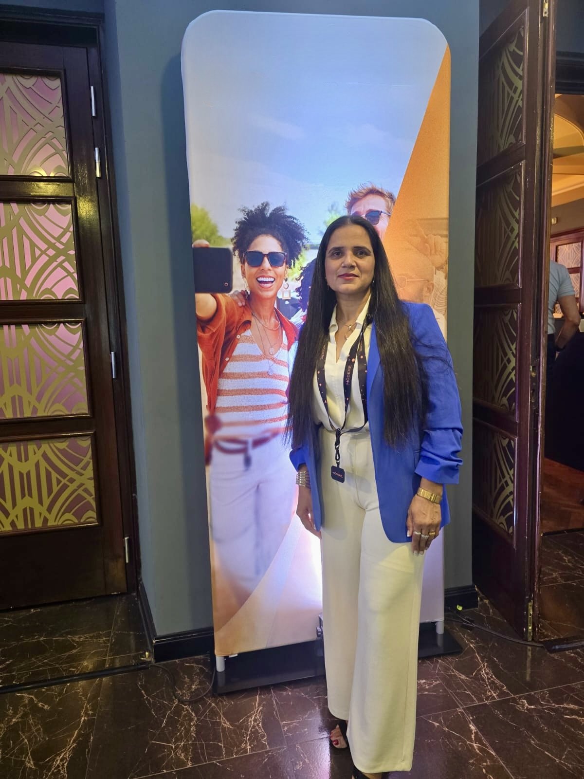 A woman with long dark hair wearing a blue blazer, white pants, and jewelry standing in front of a large vertical poster of people taking a selfie.