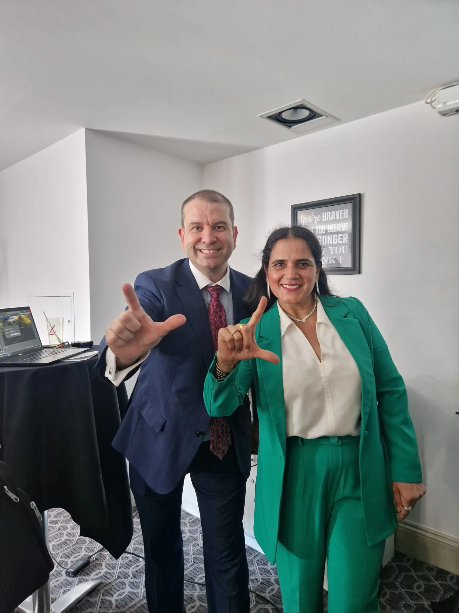A man and woman smiling and making an 'L' shape with their hands in a room with white walls, a framed quote, and a table with a laptop in the background.