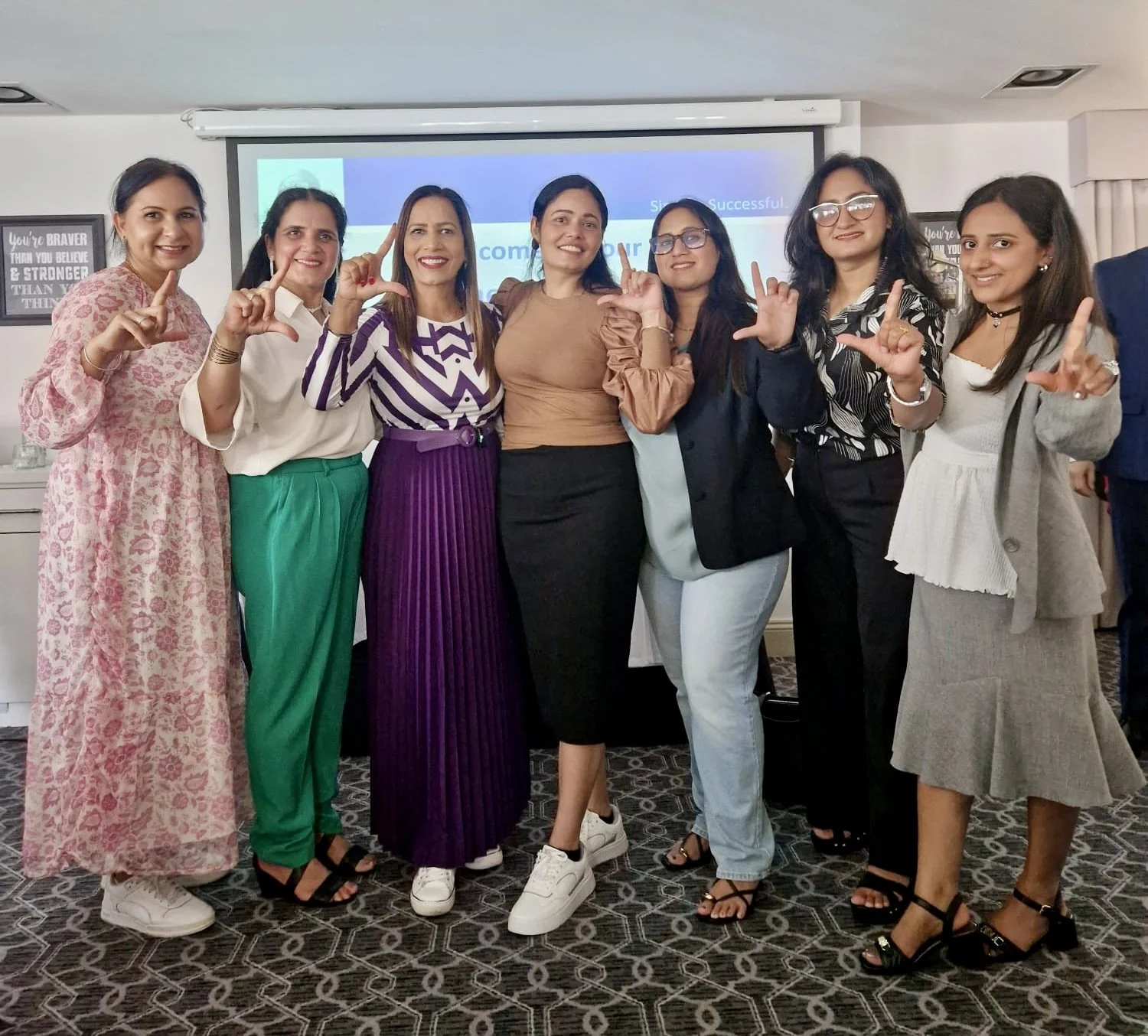 Group of women standing together in a conference room, smiling, making the 'L' sign with their hands, with a presentation screen in the background.
