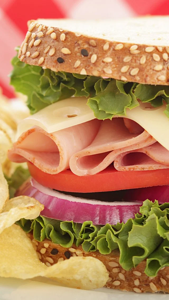 Close-up of a sandwich featuring sesame seed bread, lettuce, cheese, ham, tomato, and red onion, with potato chips beside it.
