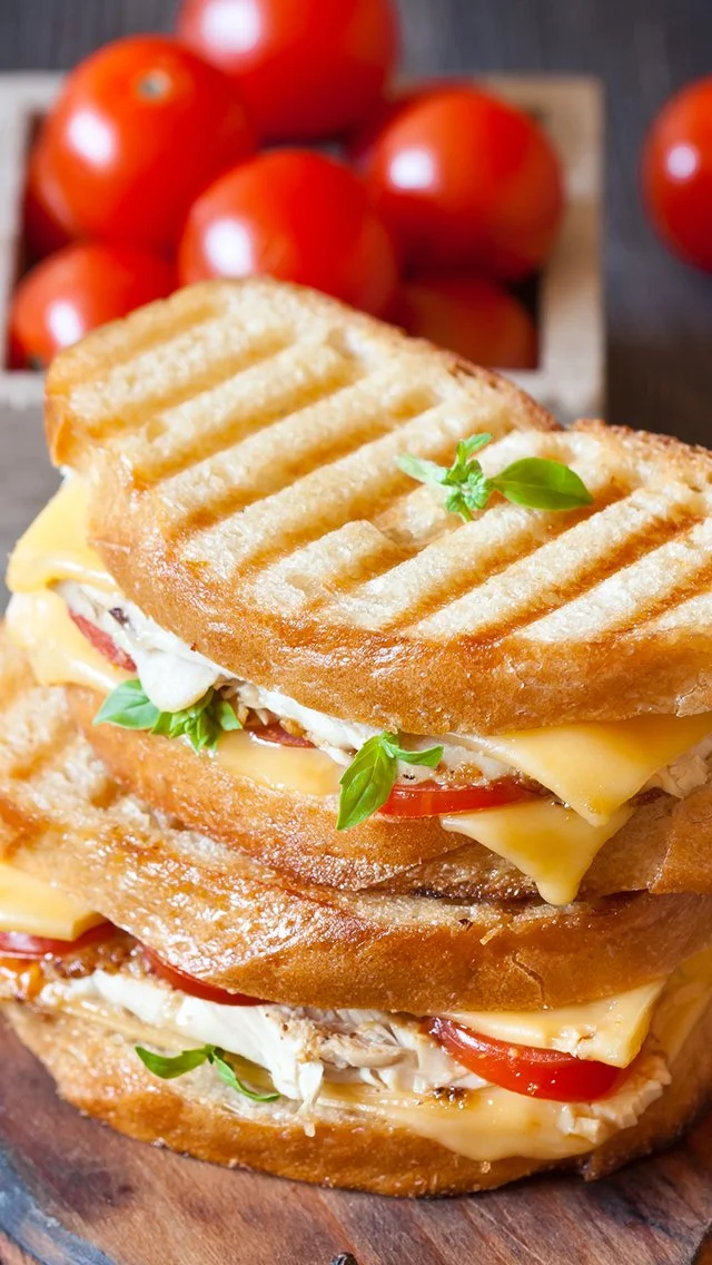 Close-up of a grilled panini sandwich with melted cheese, tomato slices, basil leaves, and grilled bread on a wooden cutting board with cherry tomatoes in the background.