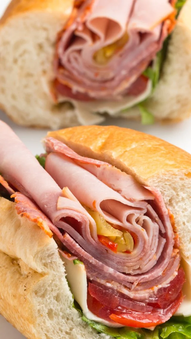 Close-up of a deli-style sandwich with sliced turkey, ham, roast beef, cheese, lettuce, tomato, and pickles in a toasted baguette.